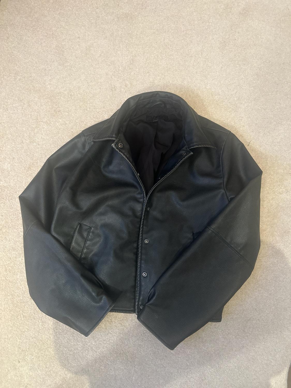 Black jacket with collar and front press-stud buttons, laid out on a light beige-toned carpet.