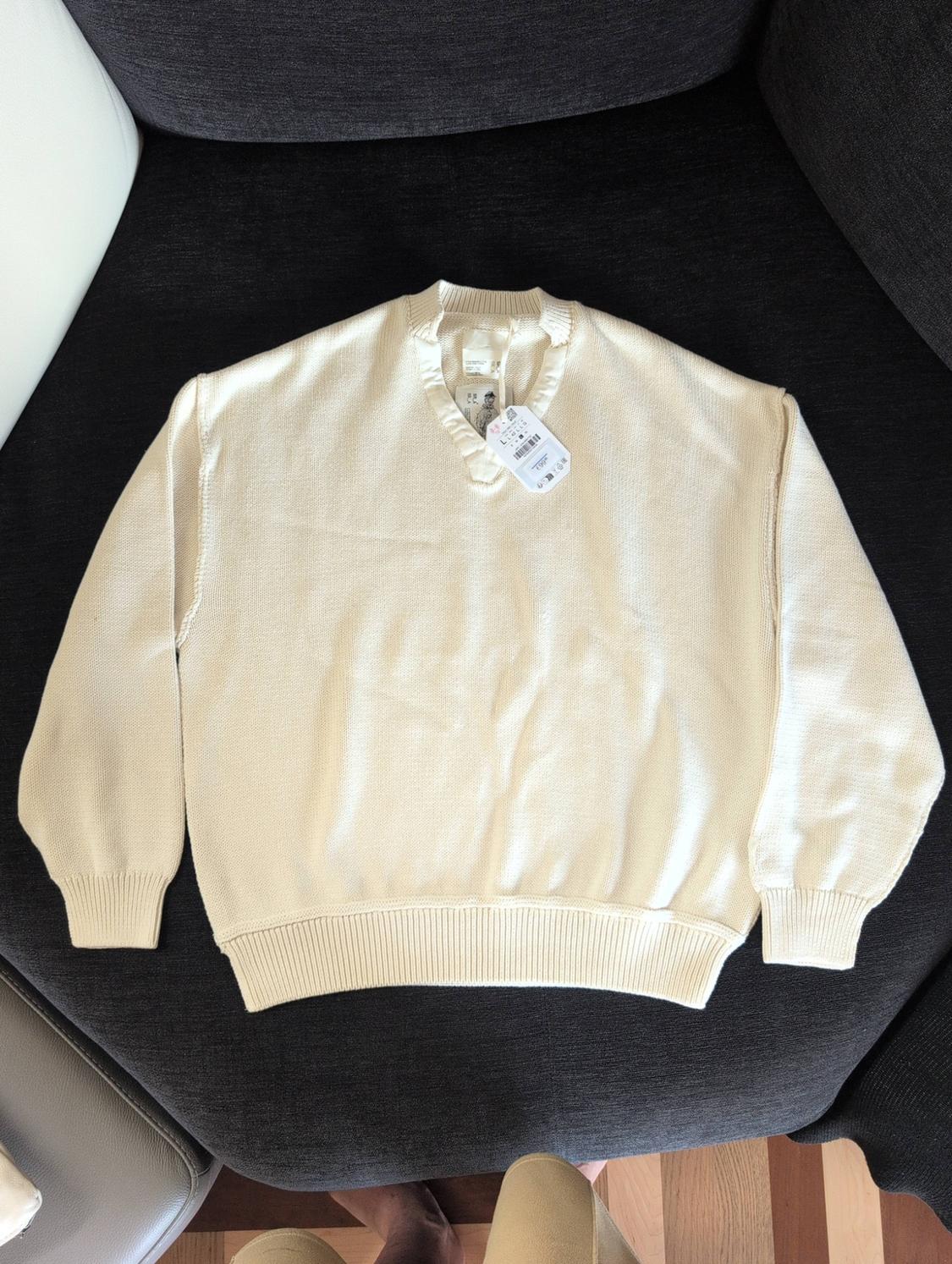 Cream-coloured knitted jumper with a V-neck and labels, laid out on a dark grey sofa.