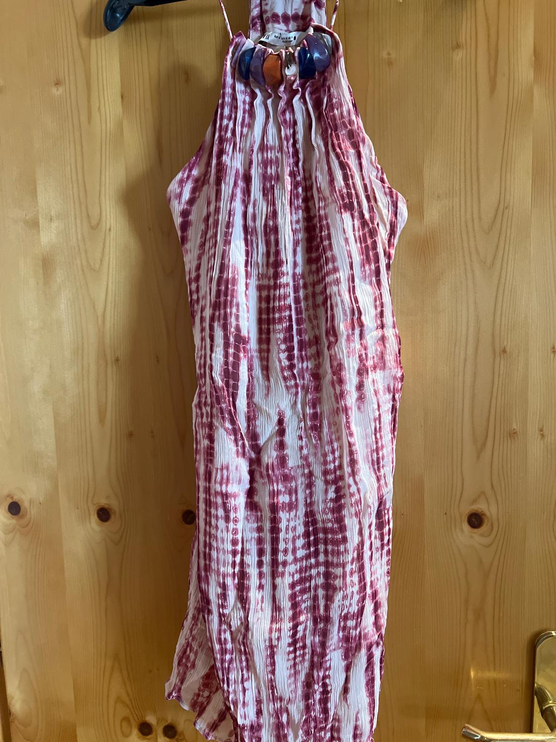 Halter-neck pleated dress with a white and maroon print and beadwork on the neckline, hanging on a hanger on a wooden door.