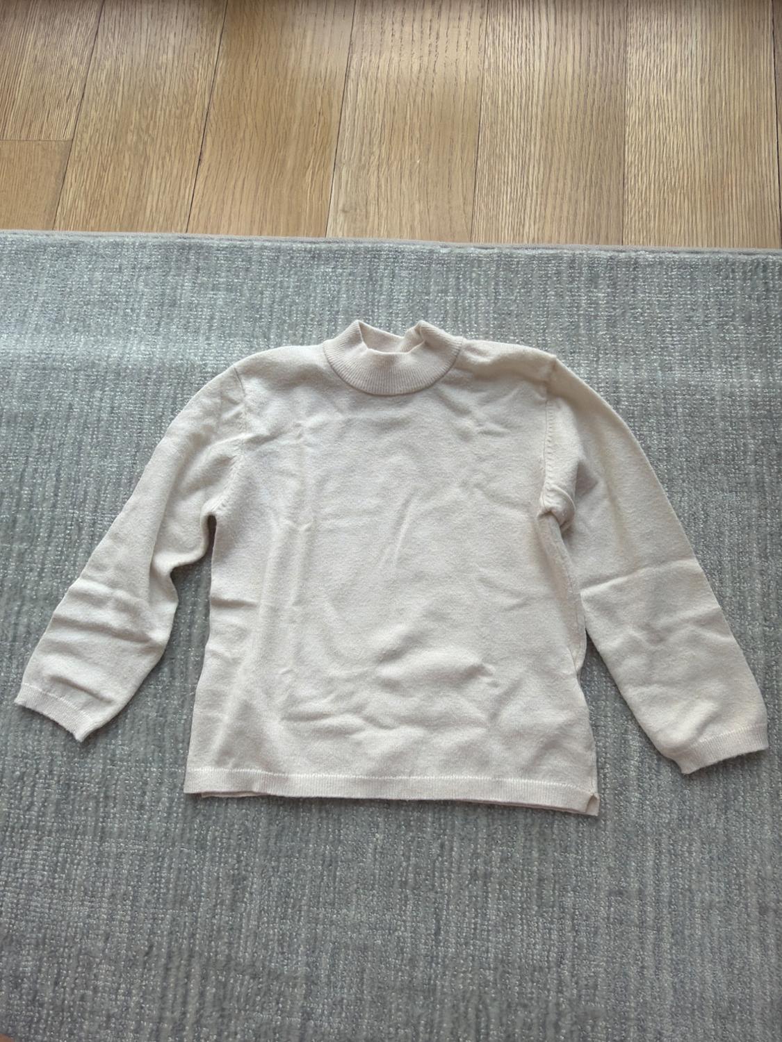 Beige roll-neck jumper on a grey rug, with a light wooden floor background.