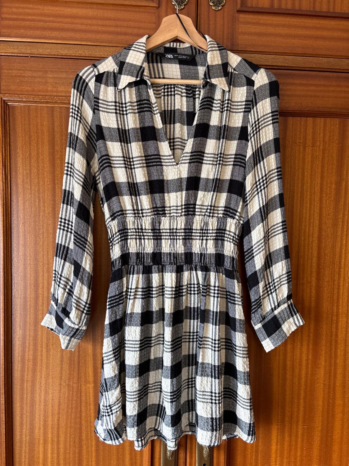 Black and white checked dress with a V-neck and long sleeves, hanging on a wooden hanger in front of a brown wardrobe.