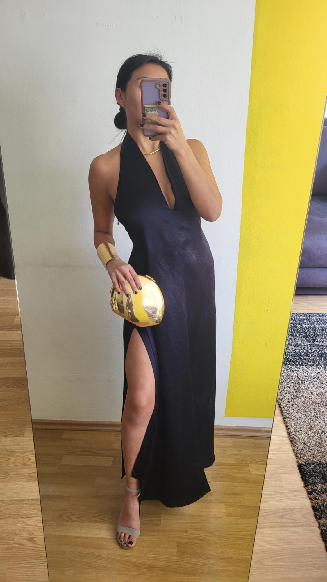 Woman wearing a long black halter neck dress with a side split, beige sandals, a gold bracelet and a gold metallic handbag in front of a mirror.