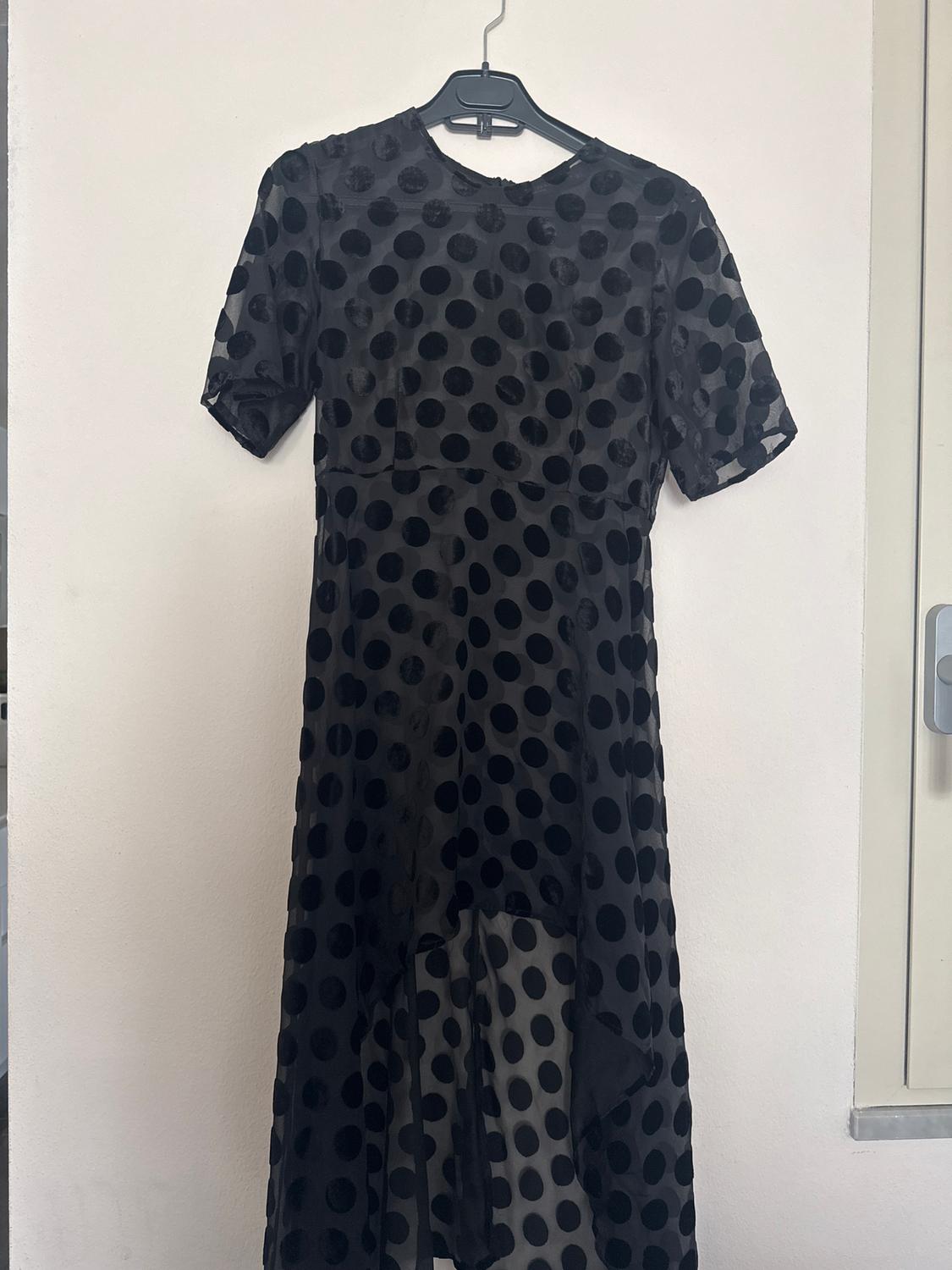 Black dress with large polka dots hanging on a hanger against a white wall. Short sleeve and asymmetrical design.