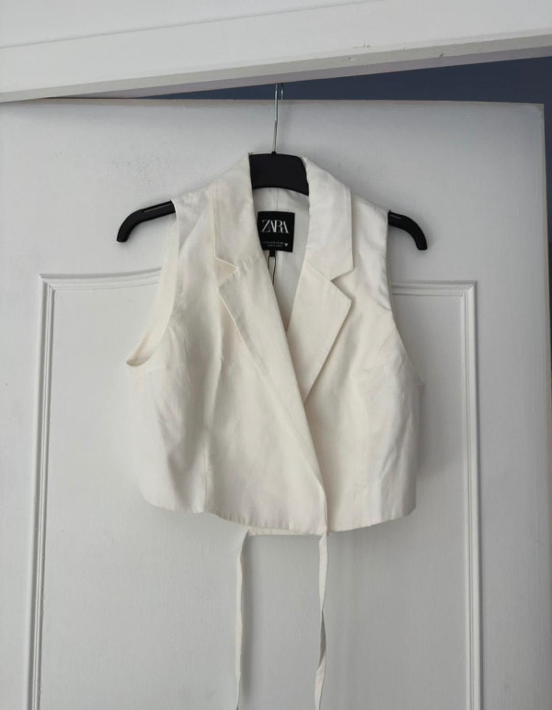 White sleeveless waistcoat hanging on a black hanger over a white door. Visible label with the text "ZARA".