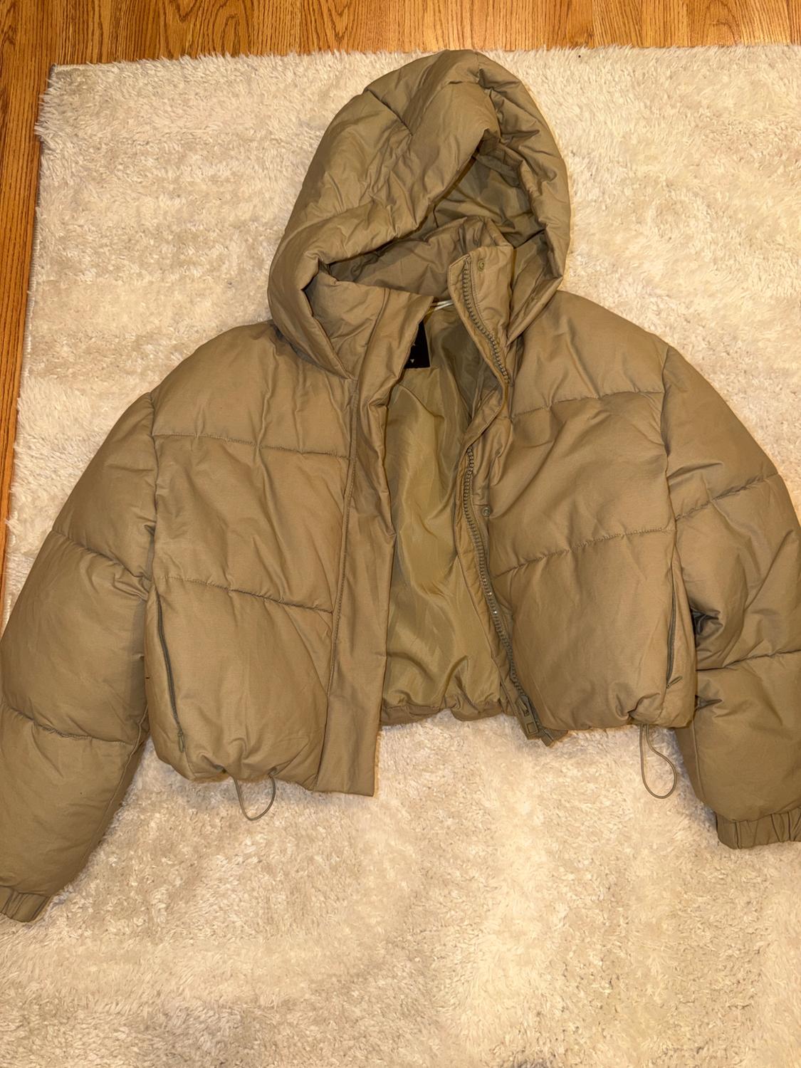 Short beige padded jacket with a hood and zip, on a fluffy white rug and wooden floor. Casual style.
