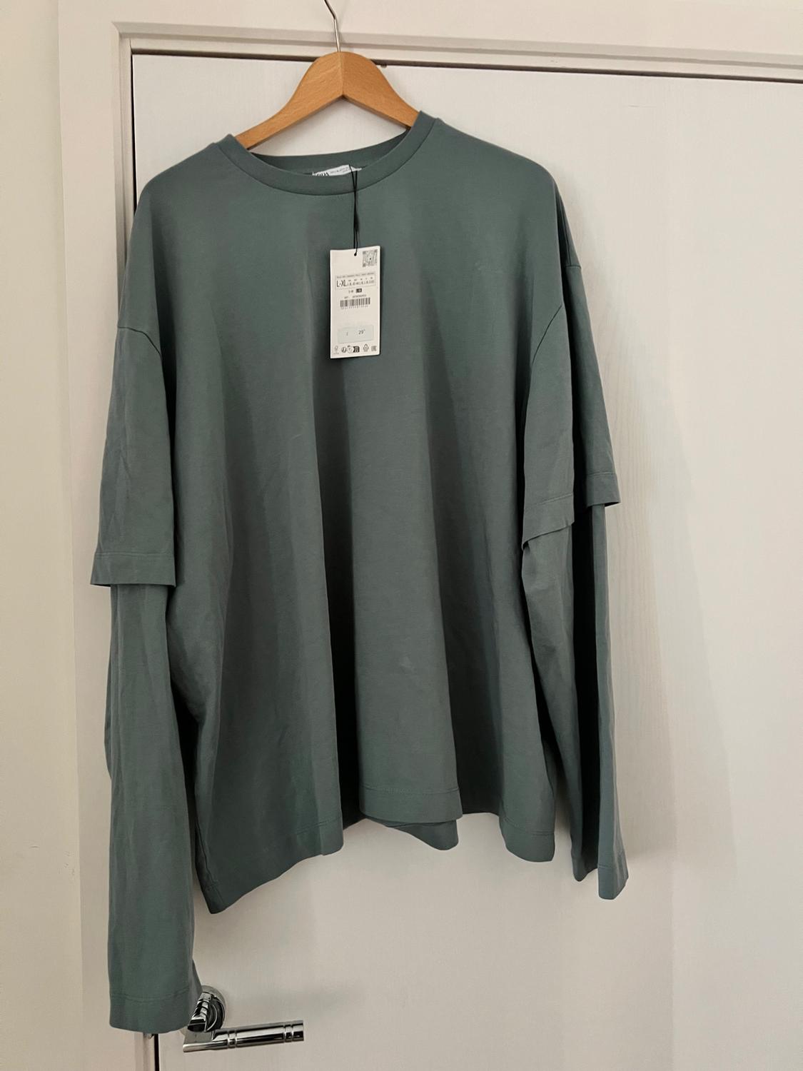Greyish-green long-sleeved T-shirt with a double-sleeve effect and label, hanging on a wooden hanger on a white door.