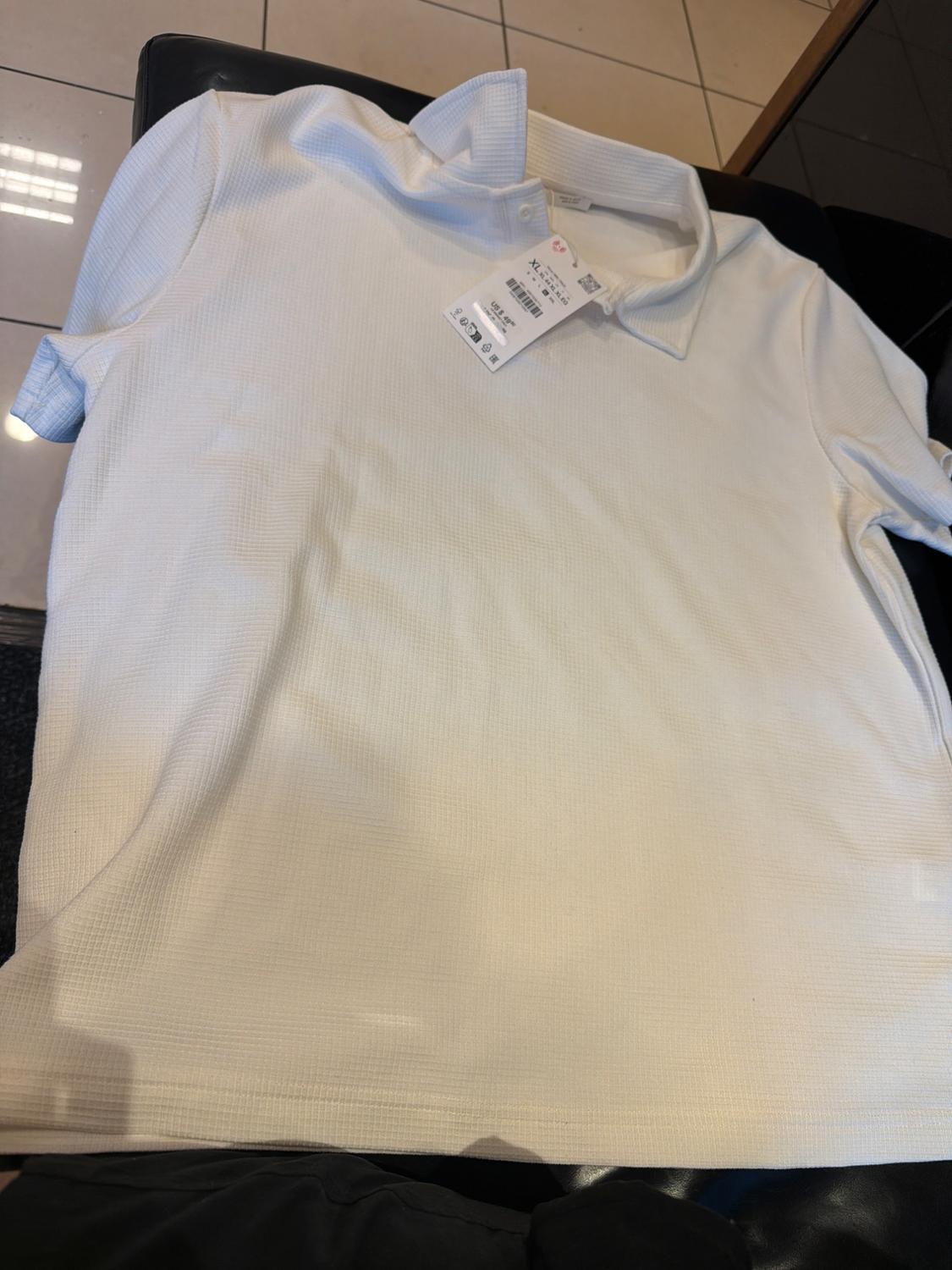 White, short-sleeved polo shirt with texture on a black surface. Label with XL text. Light tiled background. Casual and relaxed style.