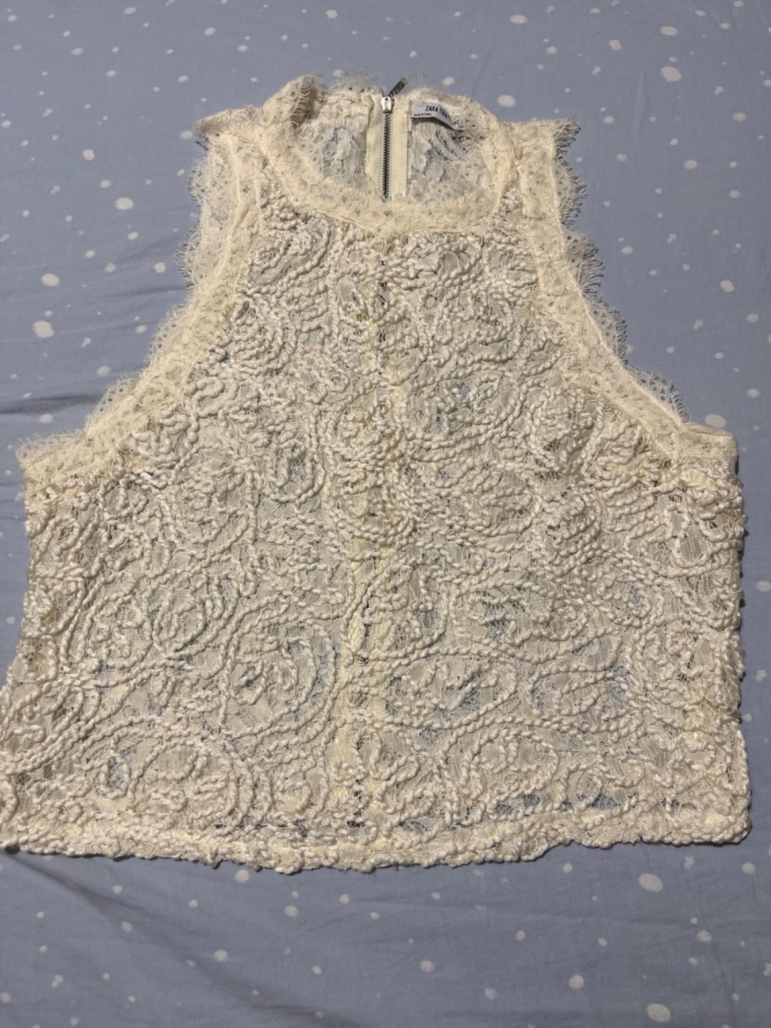 White lace sleeveless high-neck top with a back zip, set against a light blue background with white polka dots.