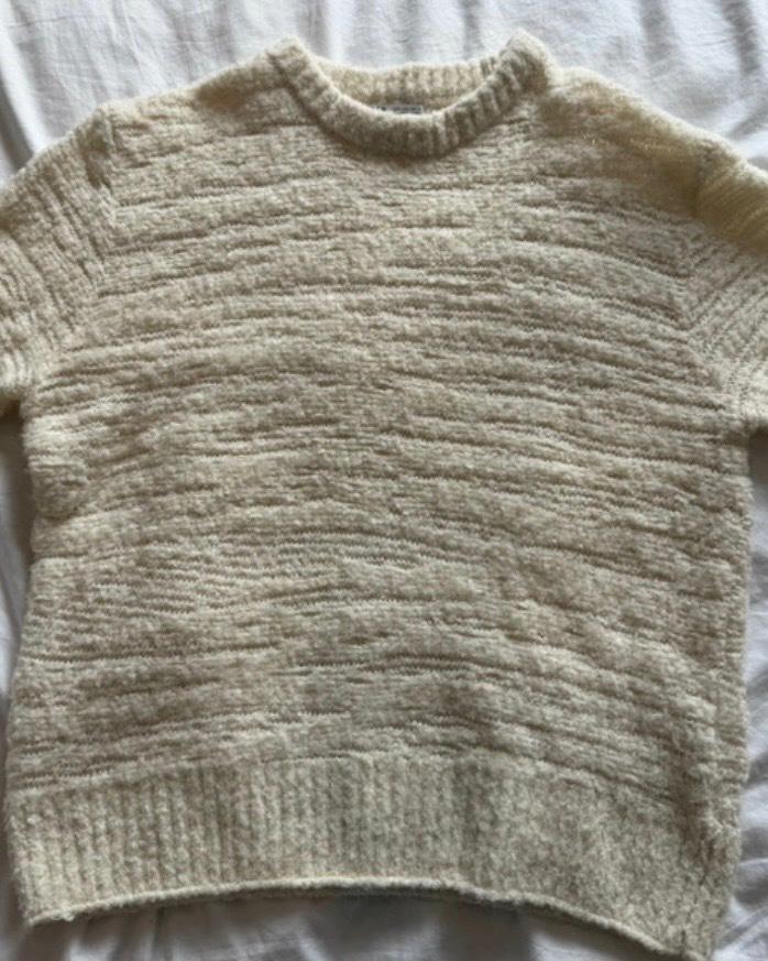 Cream short-sleeved, round-neck knit jumper with a horizontal line texture, laid on a crumpled white sheet. Casual style.