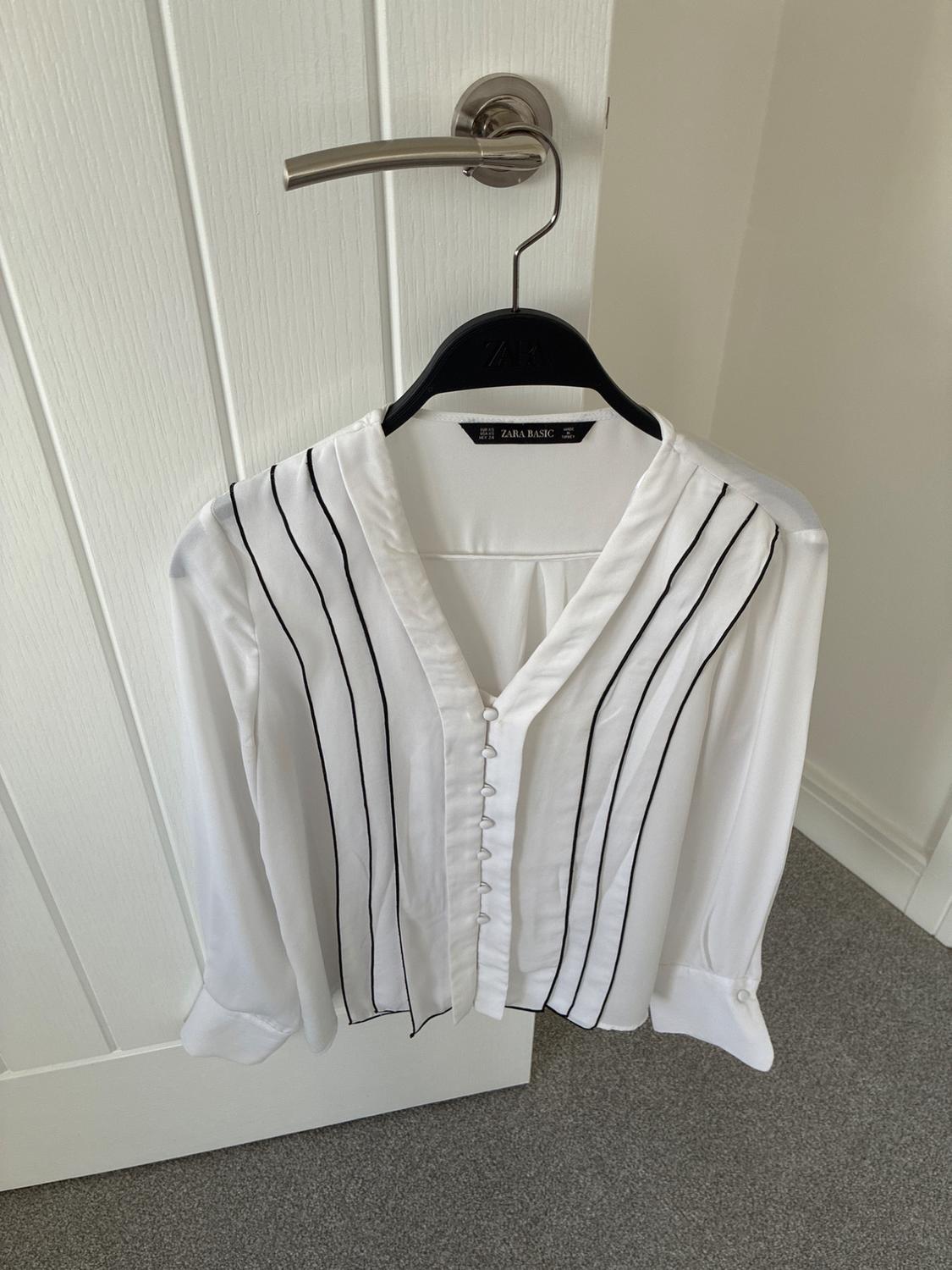 White long-sleeved blouse with black stripes and buttons, hanging on a white door. Text ZARA BASIC on the label.