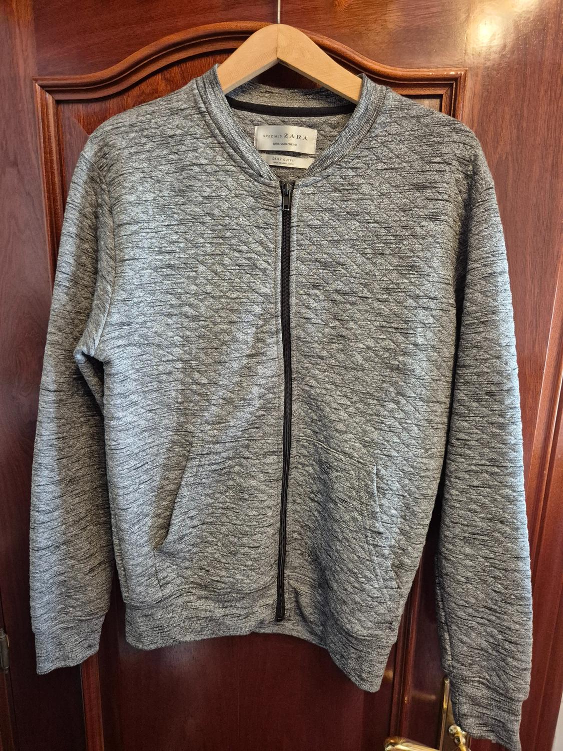 Grey quilted zip-up jacket on wooden door. Text: SPECIALS ZARA EUR M / USA M / MEX 40 DAILY OUTFIT MADE IN BANGLADESH.