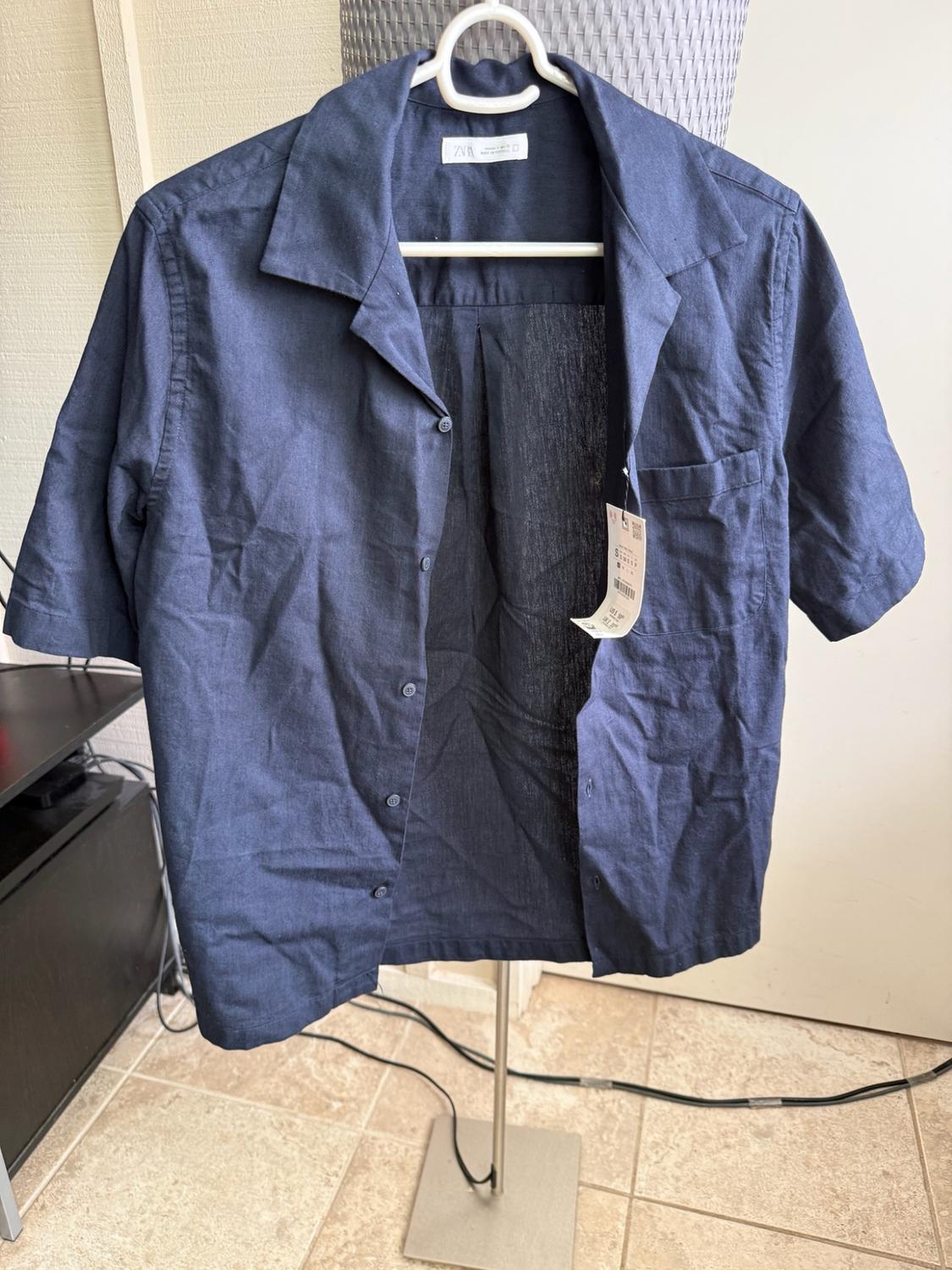 Unbuttoned navy blue short-sleeved shirt with a ZARA label, hanging on a white hanger on a metal stand.