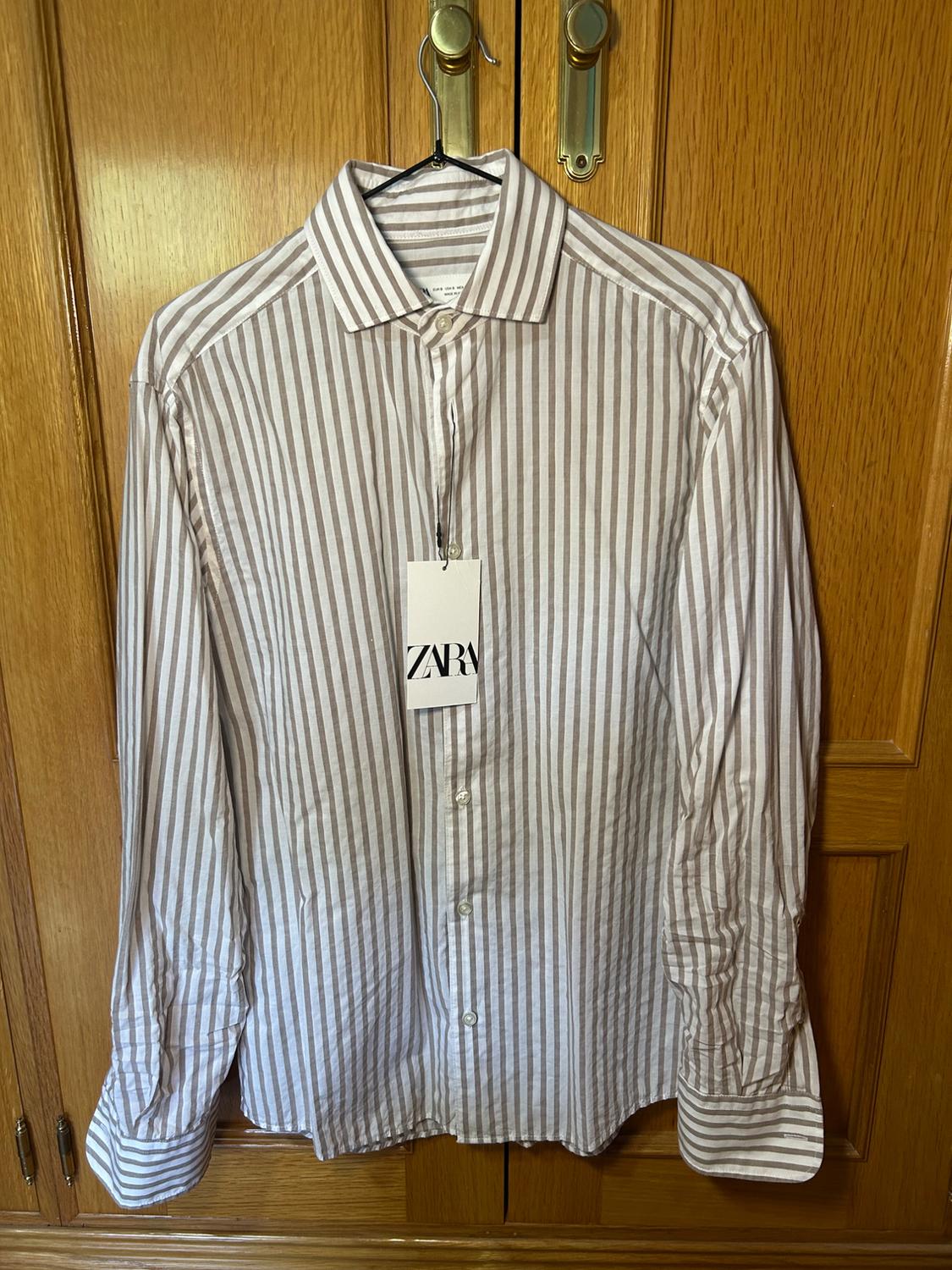 Long-sleeved shirt with white and beige stripes hanging in a wooden wardrobe. Visible label with the text ZARA.