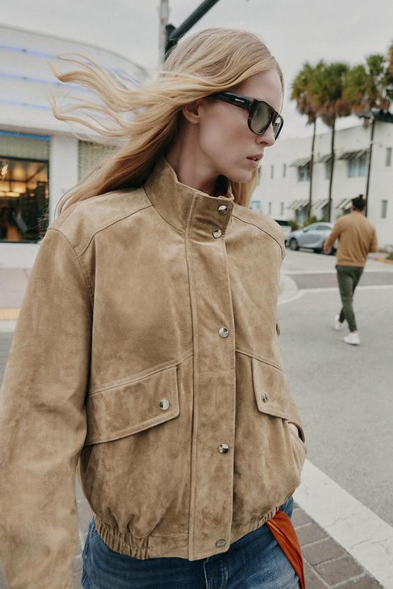 "Střední detail béžové bundy s vysokým límcem, kovovými knoflíky a předními kapsami s klopou."