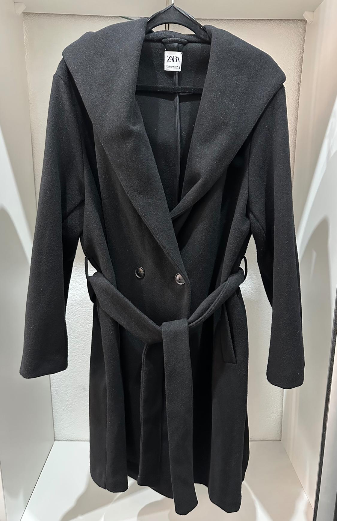 Black coat with a hood and belt, hanging on a hanger. Visible label with the text "ZARA". White background.