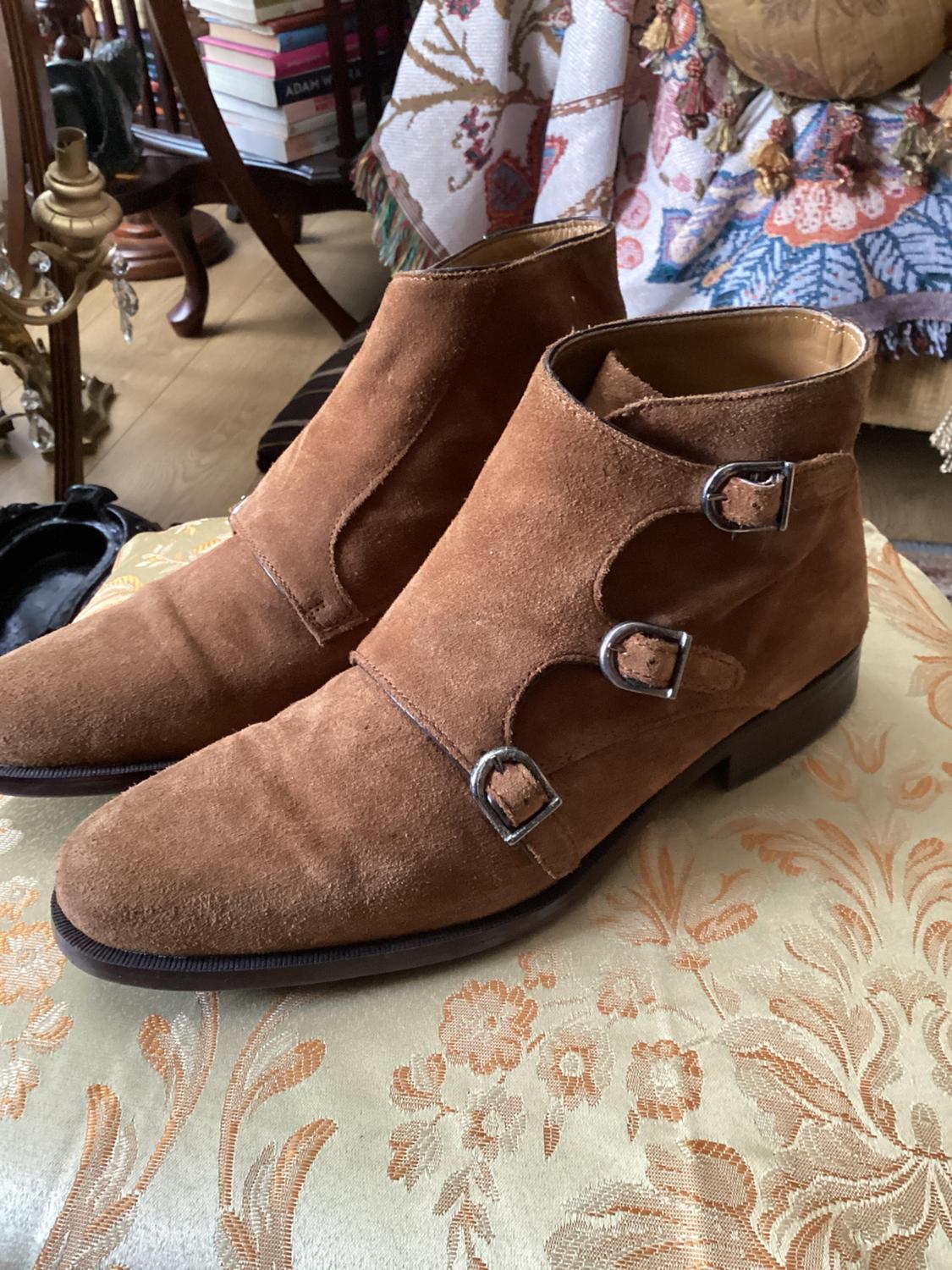Brown suede boots with metal buckles on a floral-patterned cushion. Background with books and classic decor.