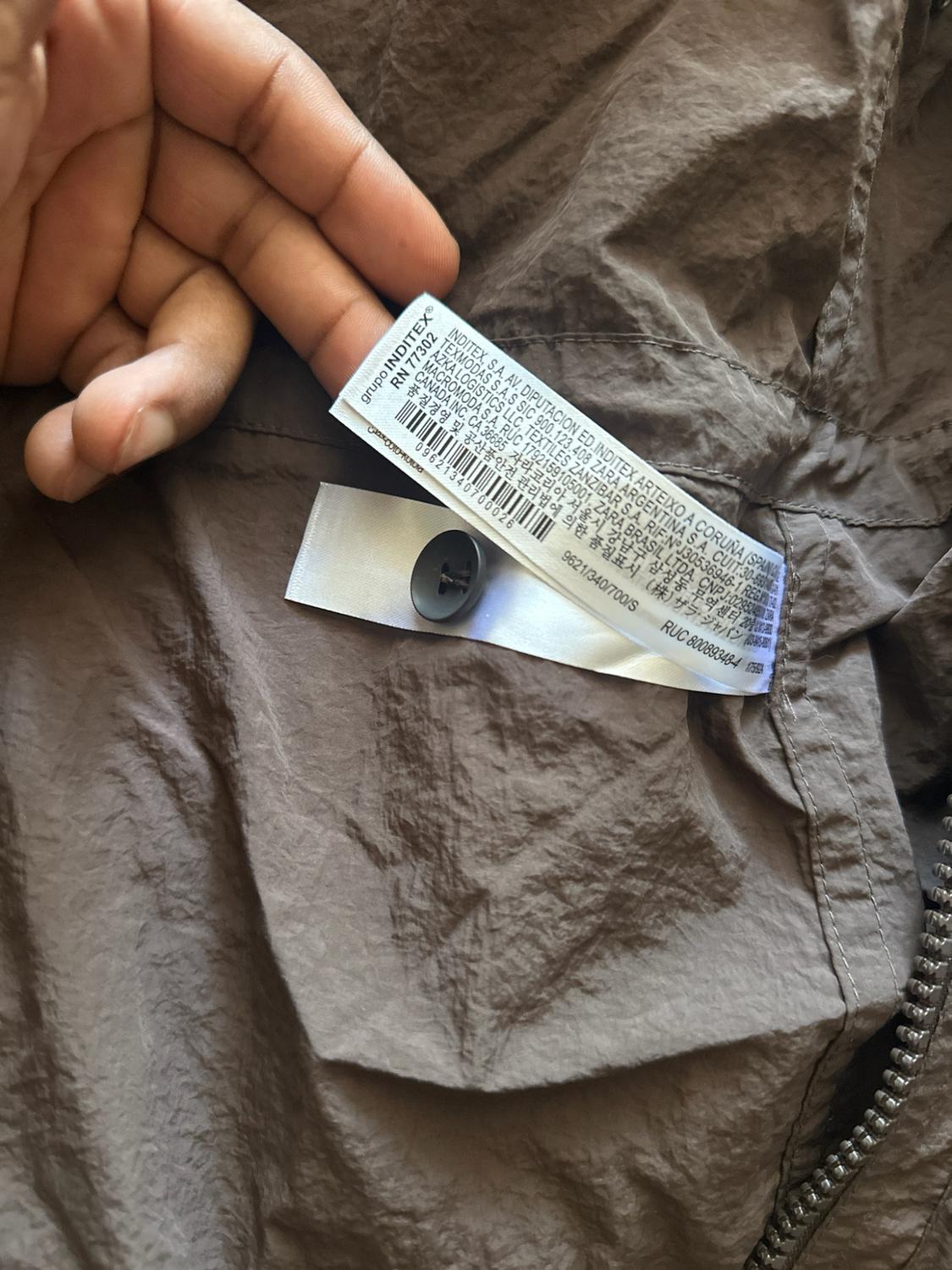 I’m holding tags with the text group INDITEX RN 77302 and a spare button over the wrinkled brown fabric of a garment.