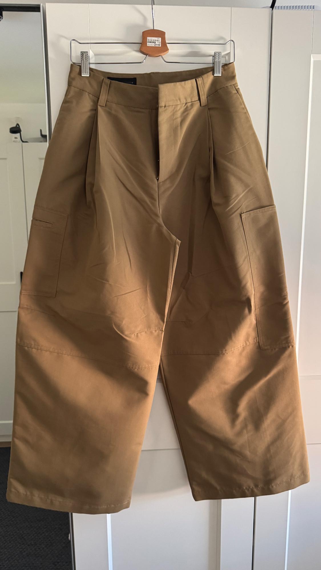 Brown shorts hanging on a metal hanger over a white door. Large pockets and pleats visible.