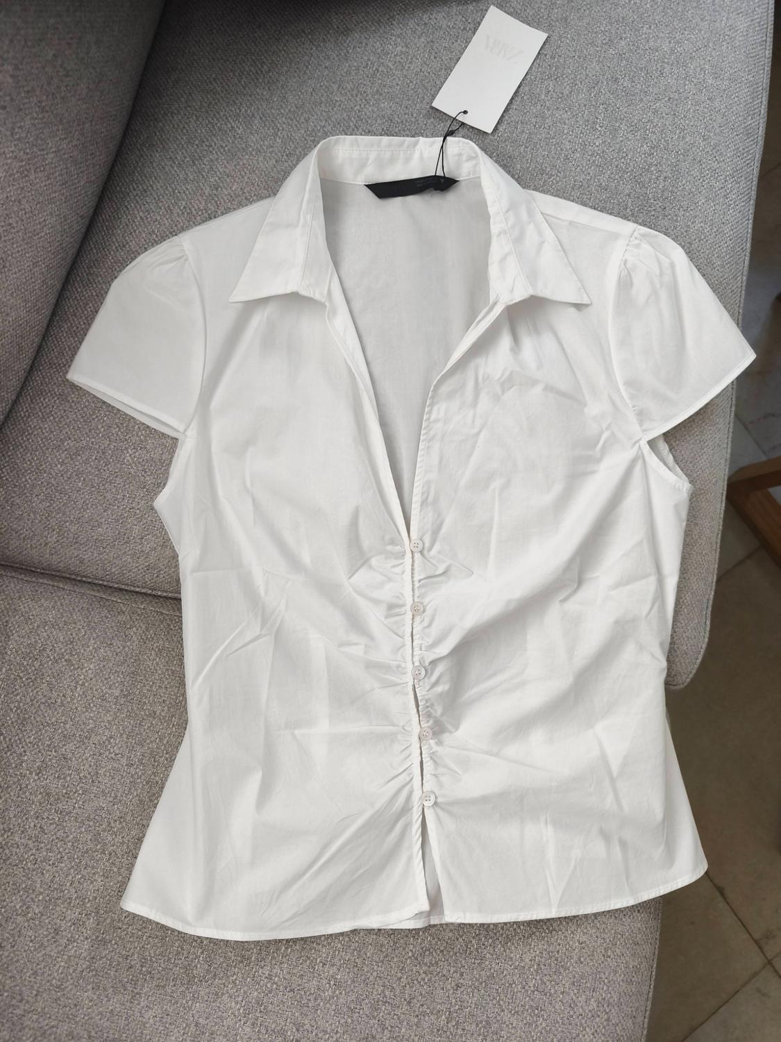 White short-sleeved shirt with a collar and gathered front buttons, on a grey sofa. Label with the text ZARA visible.