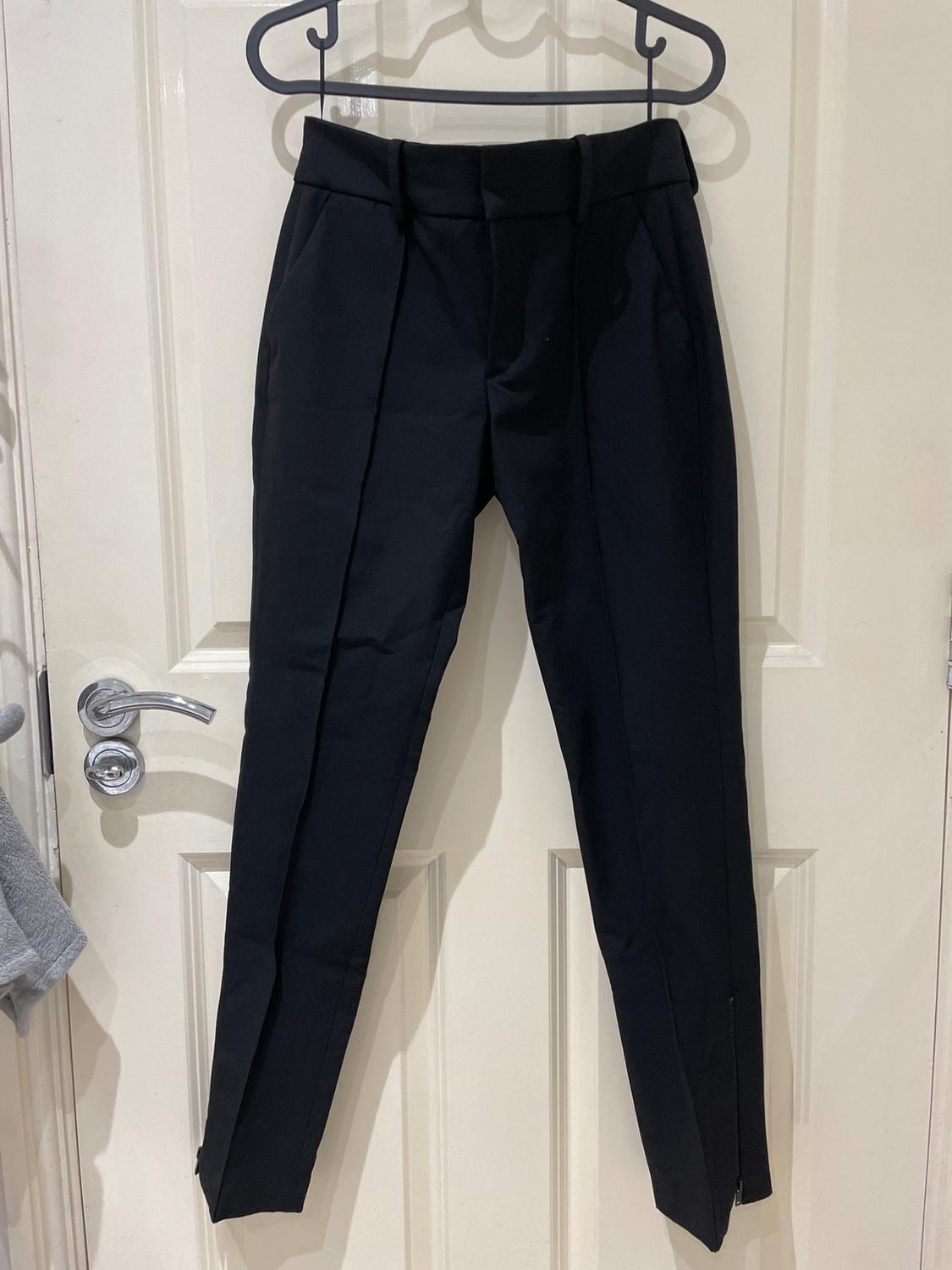 Black trousers with a front seam and zip detail at the hem, hanging on a white interior door. Casual and relaxed style.