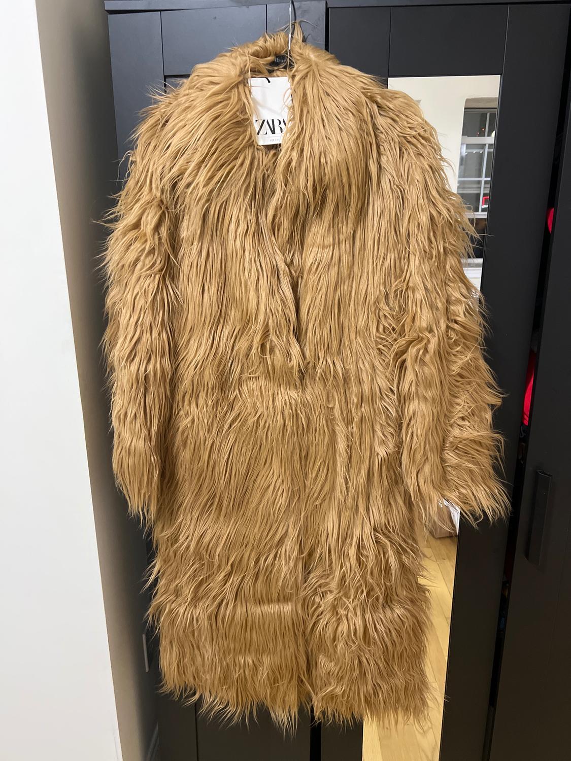 Long camel faux fur coat hanging in a black closet. Visible tag with "ZARA" text. Interior room background.