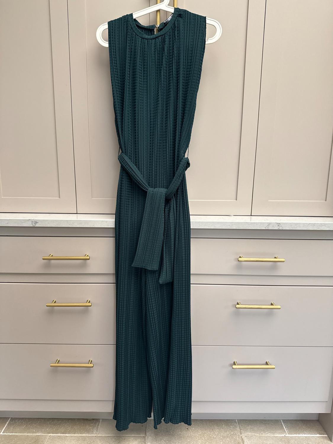 Dark green sleeveless pleated jumpsuit with a belt, hanging on a hanger in front of beige cupboards with gold handles.