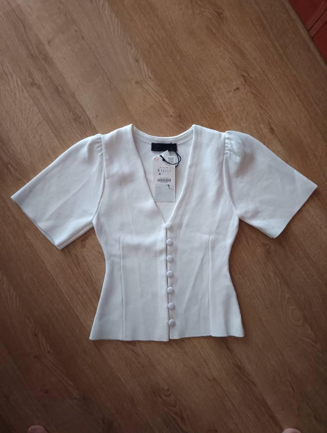 White top with short puff sleeves, a V-neck and front buttons, on a wooden floor.