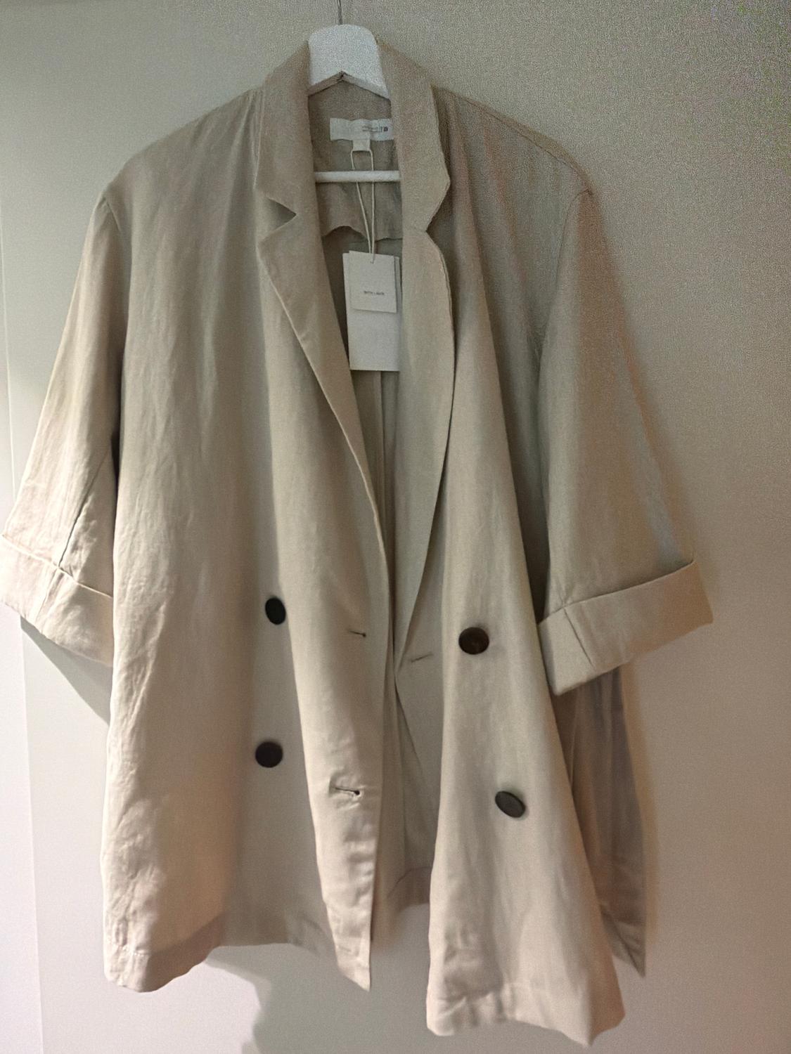 Beige trench coat hanging on a white hanger, with dark buttons and a visible label. Neutral background.