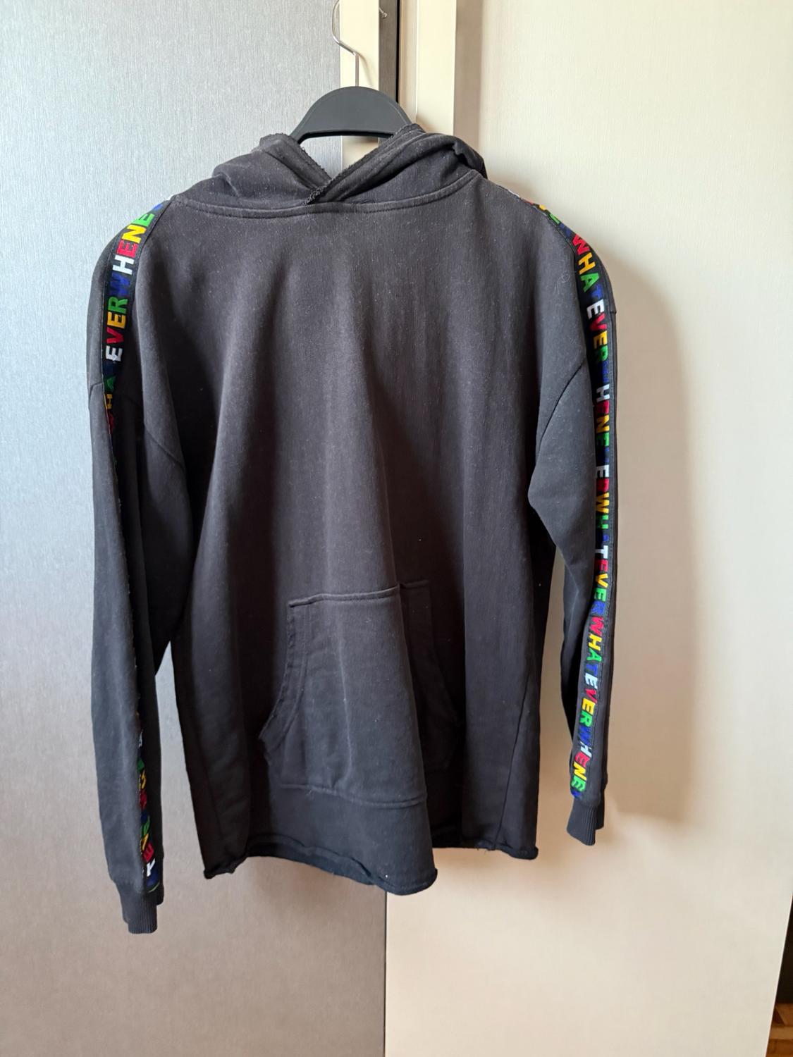 Black hooded sweatshirt hanging on a hanger. Front pocket and bands on the sleeves with the multicoloured text "WHATEVER WHENEVER".