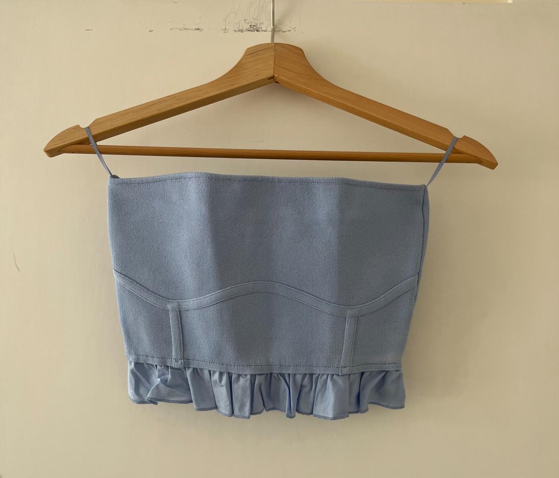 Light blue camisole top with thin straps, stitching and ruffled hem, hanging on a wooden hanger against a light-coloured wall.