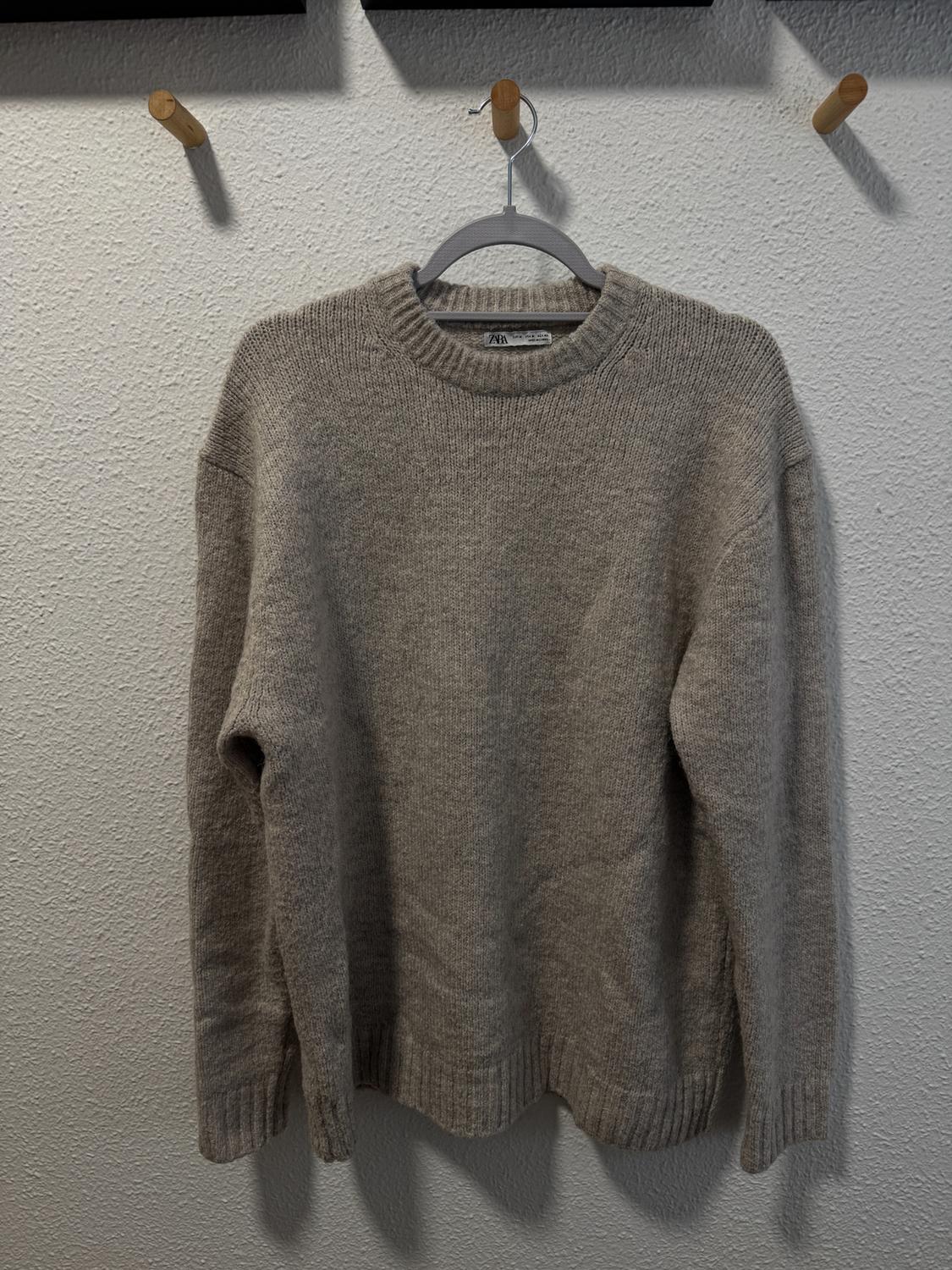 Chunky beige knitted jumper hanging on a grey hanger against a textured white wall.