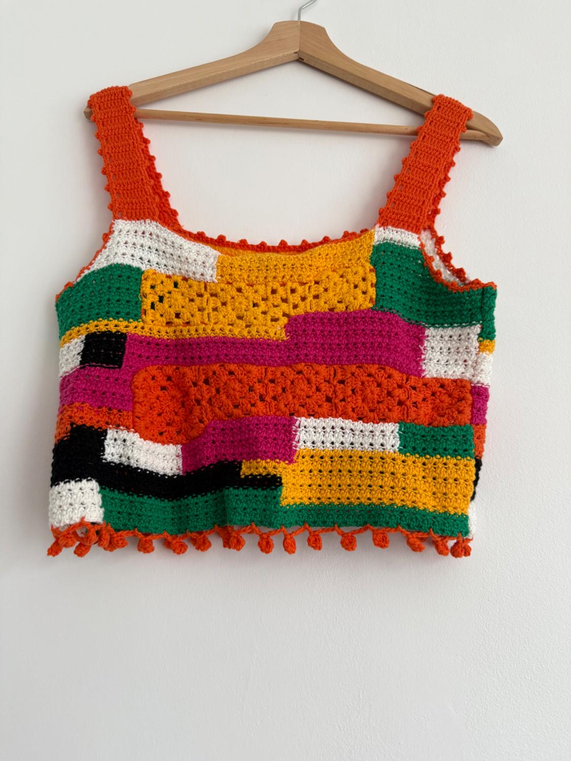 Multicoloured crochet top with wide straps, hanging on a wooden coat hanger against a white wall.