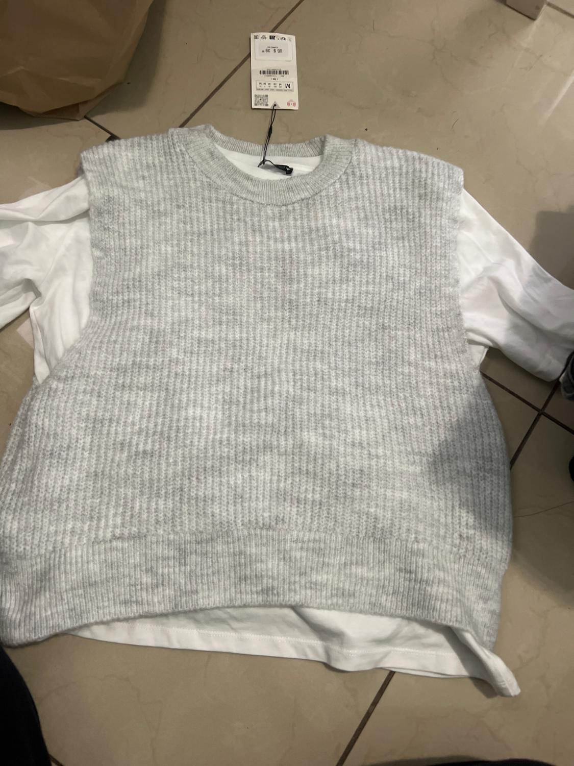 Grey knitted waistcoat over a white t-shirt, with a visible price tag, placed on a beige tiled floor.
