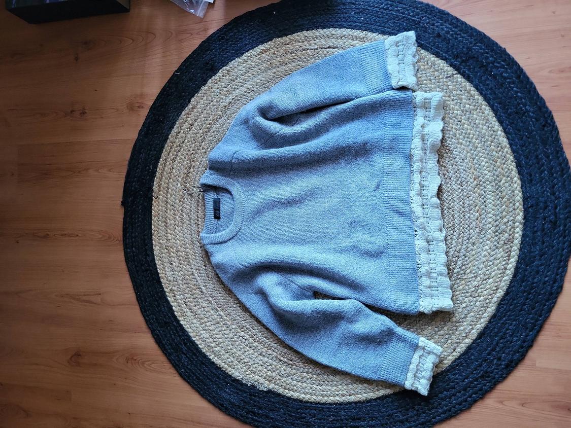 Grey jumper with lace details on the edges, placed on a circular jute rug on a wooden floor.