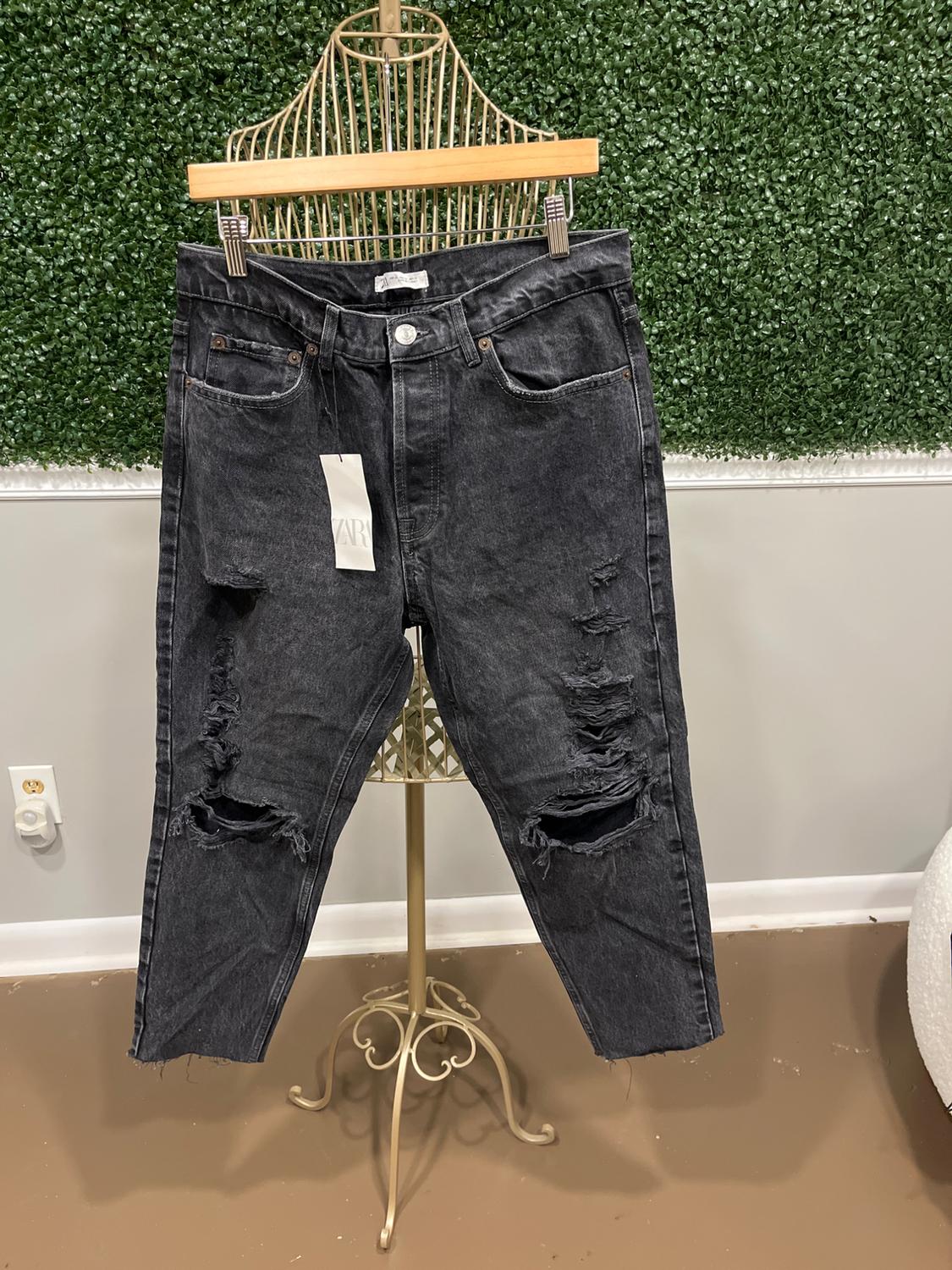 Distressed black jeans hanging on a golden metal mannequin, with an artificial leaf green wall background. Visible tag.