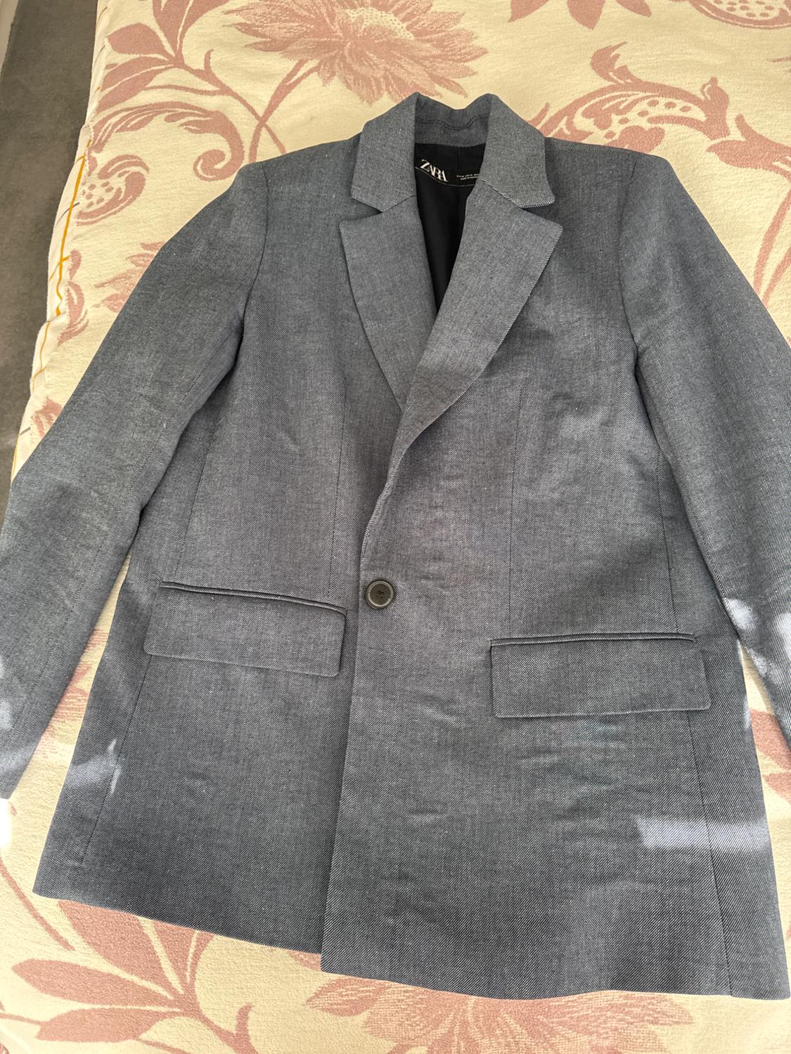 Single-button grey jacket on a floral-patterned quilt in beige and pink tones. Visible label inside.