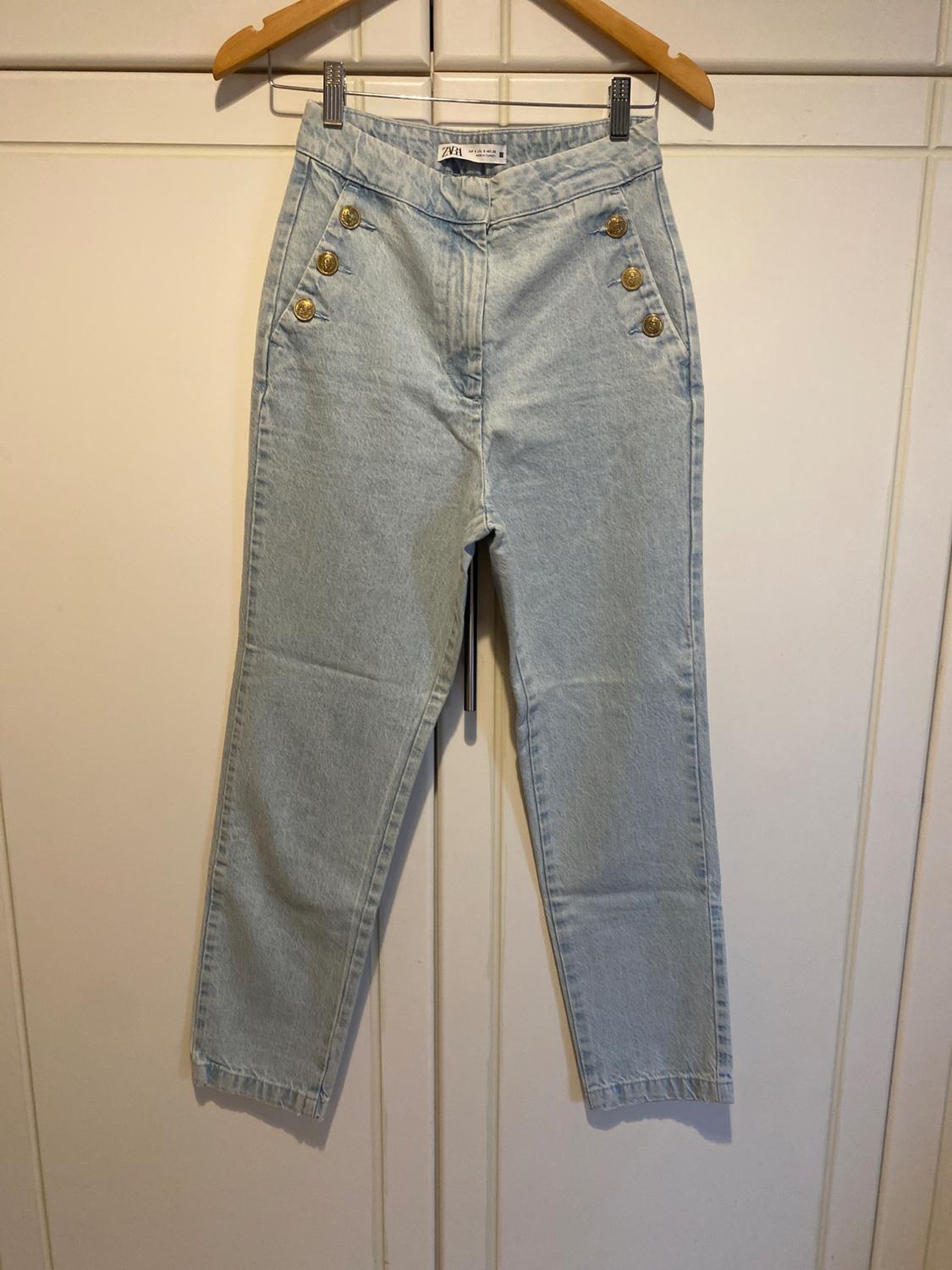 Light blue denim jeans with gold buttons, on a wooden hanger over white wardrobe doors. Label with the text ZARA.