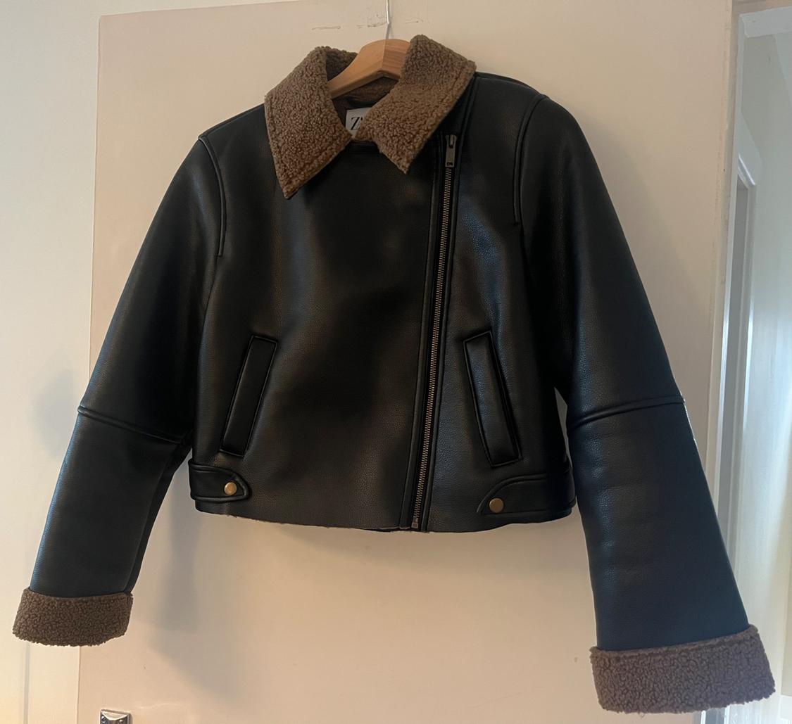 Black jacket hanging on a white door. It has an asymmetric zip, pockets, and a brown fur collar and cuffs.