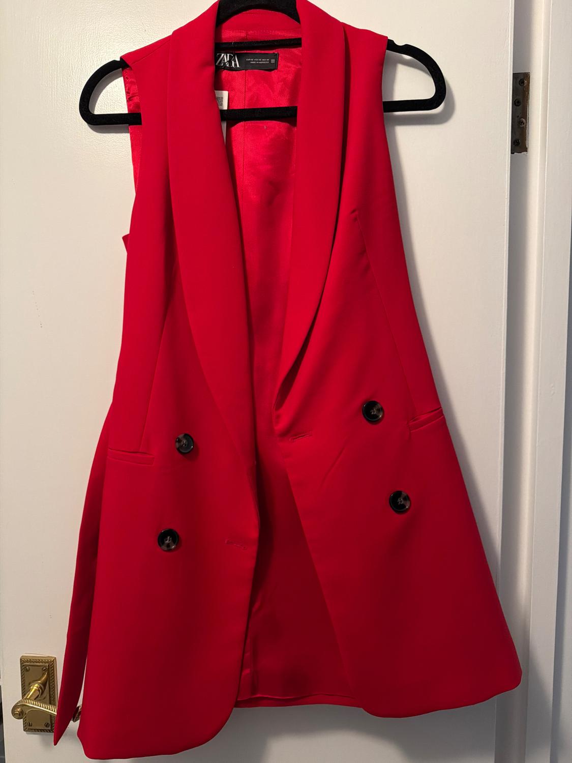 Red sleeveless waistcoat with wide lapels and black buttons, hanging on a black hanger over a white door.