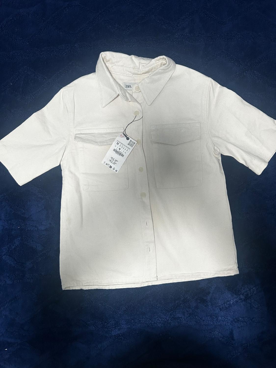 Short-sleeved white shirt with two front pockets and hangtag, on a dark blue background.