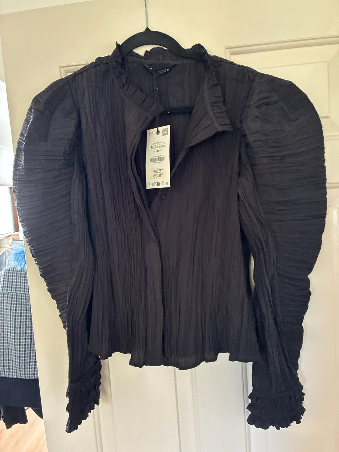 Black crinkled-texture blouse, puff sleeves and frilled cuffs,
hanging on a hanger over a white door. Label visible.