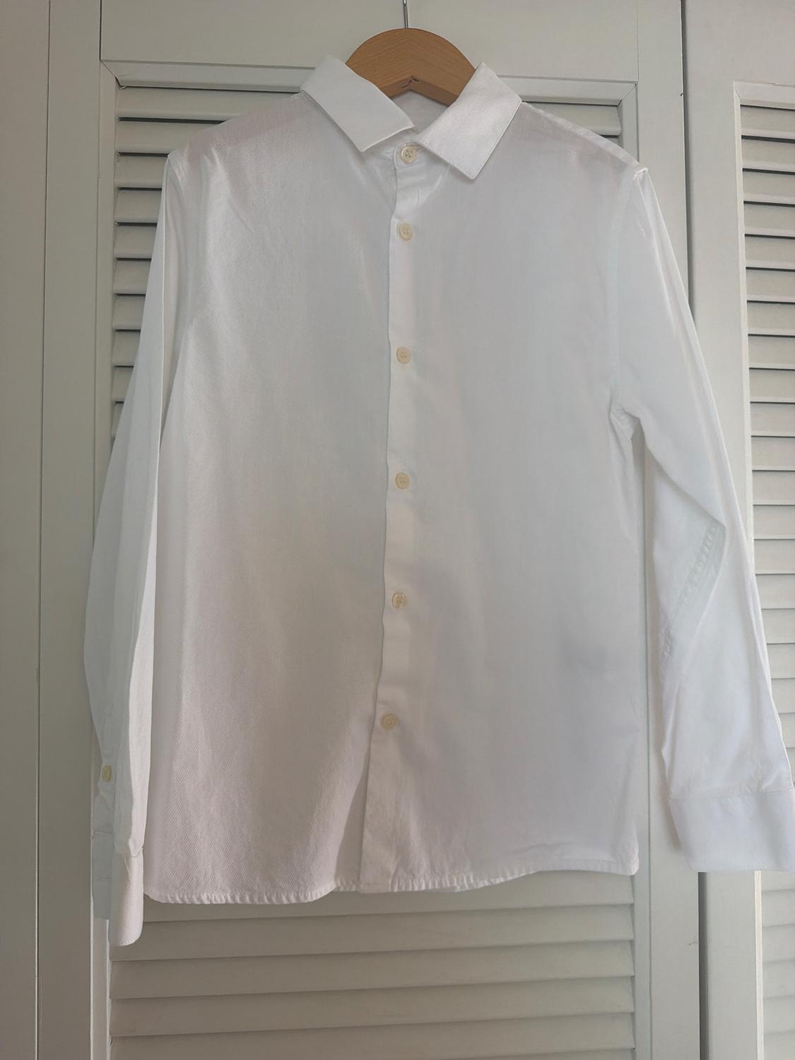 White long-sleeved shirt hanging on a wooden hanger in front of a background of louvered wardrobe doors.