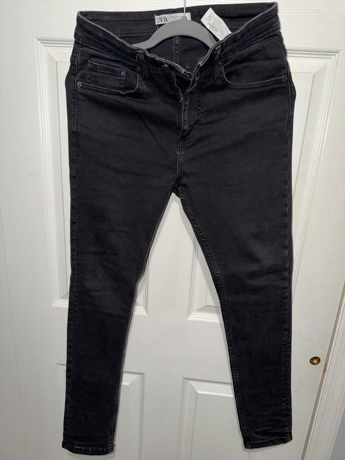Tight black trousers hanging on a hanger in front of a white door. Label visible at the waist.