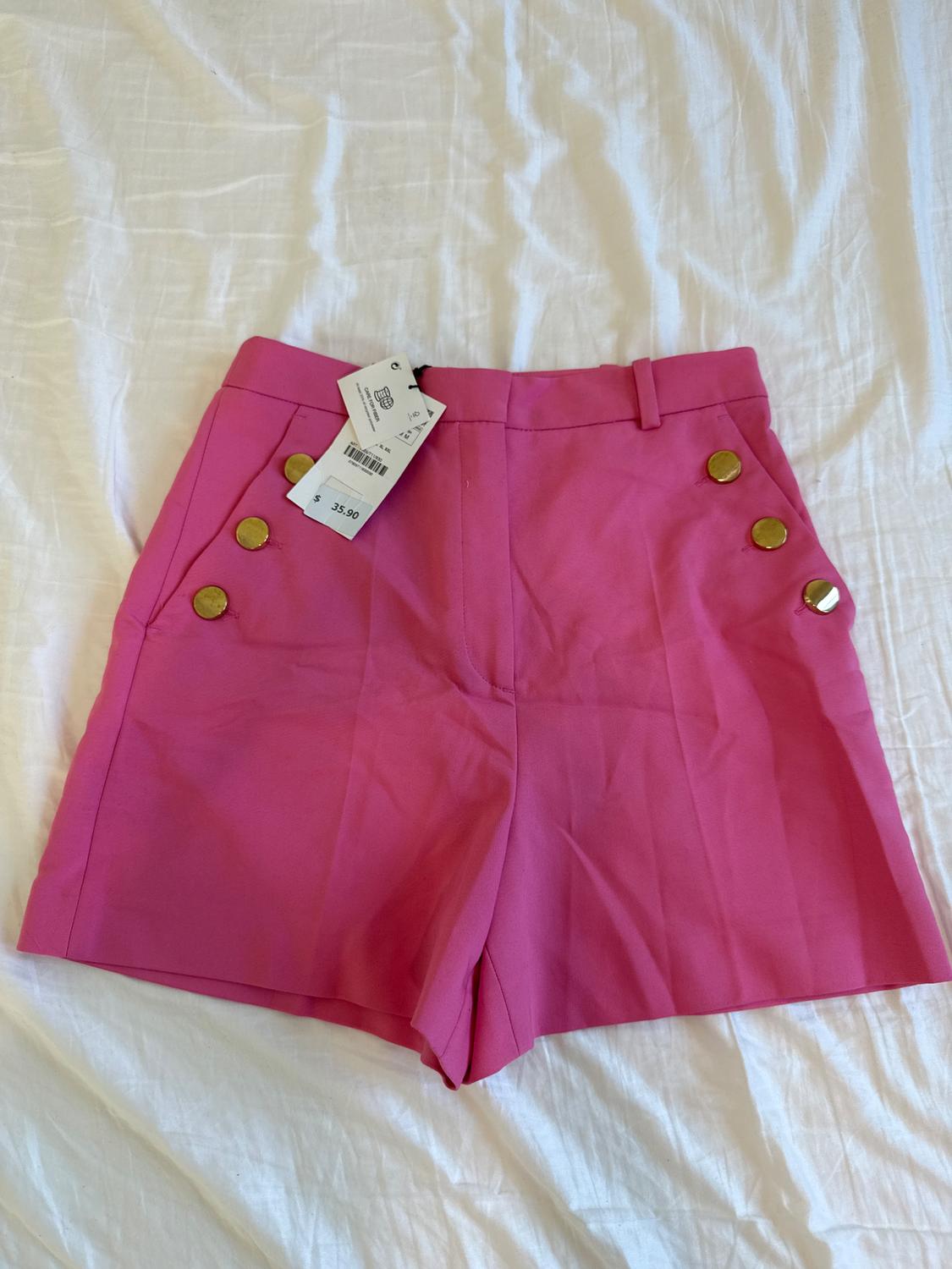 Pink shorts with gold buttons on the sides, on a white fabric. Visible tag with price of 35.90.