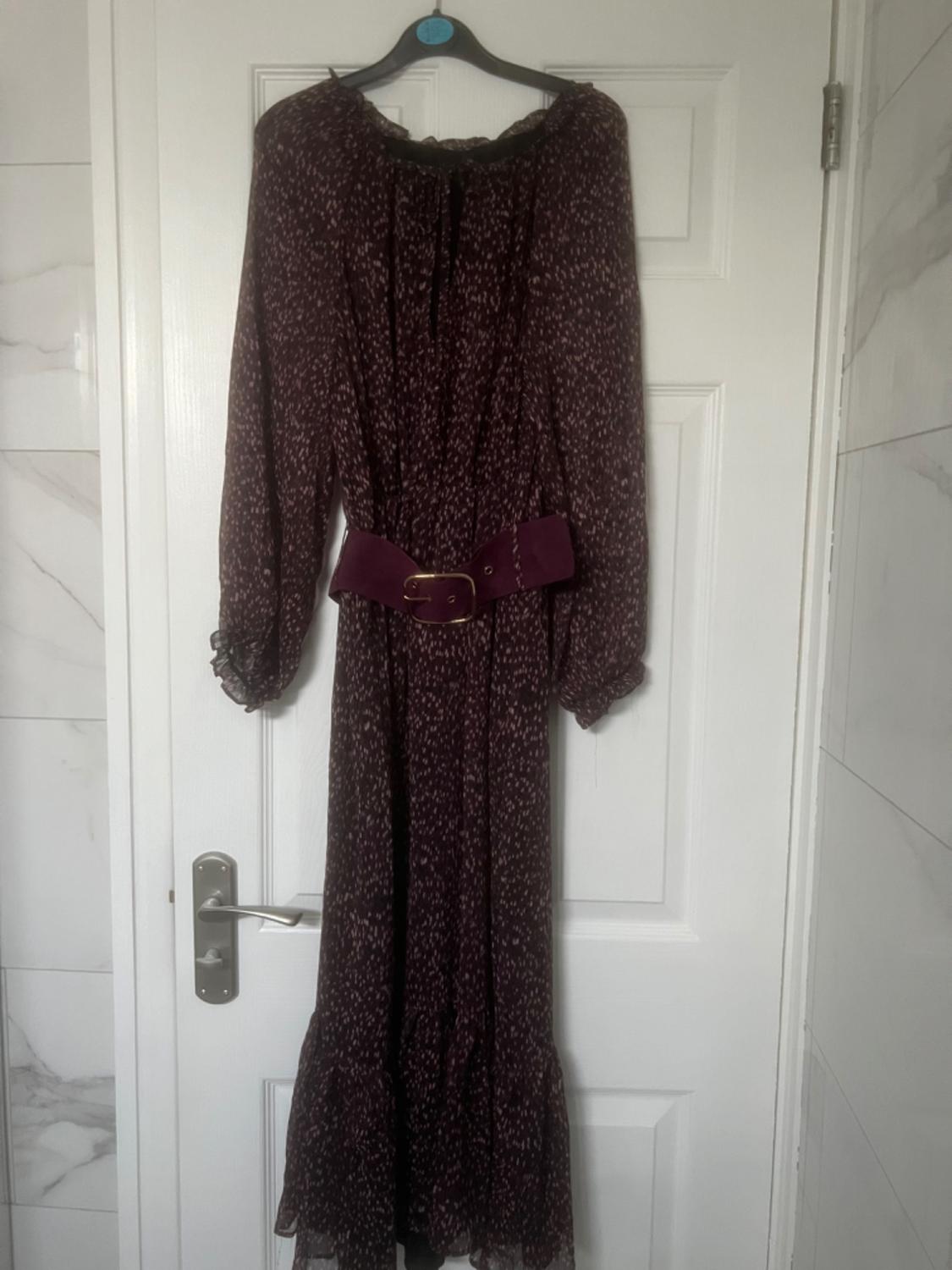 Long printed dress in dark tones with a purple belt, hanging on a white bathroom door with light tiles.