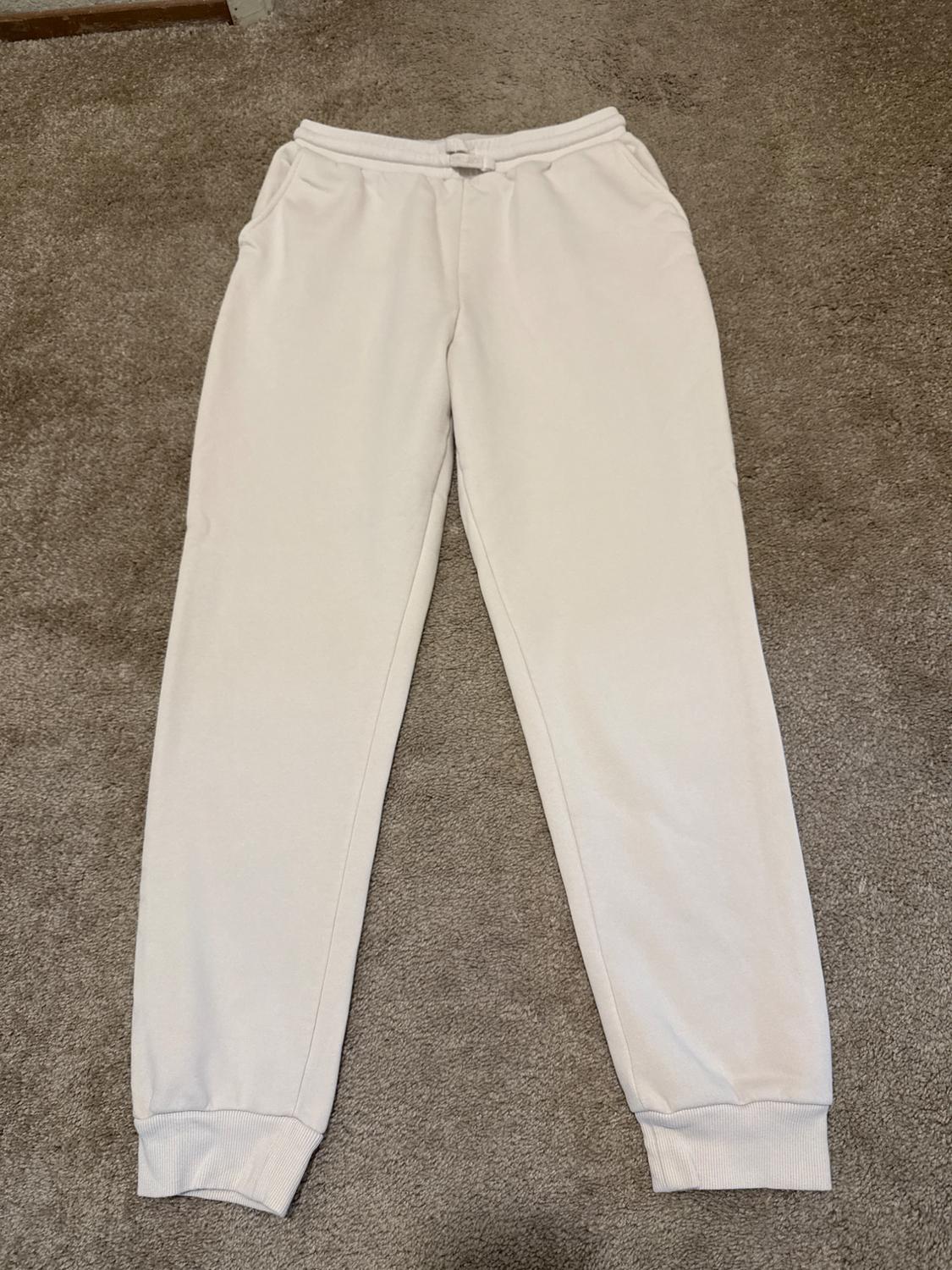 White sweatpants with an elastic waistband, drawstring, pockets, and cuffs, on a brown rug.