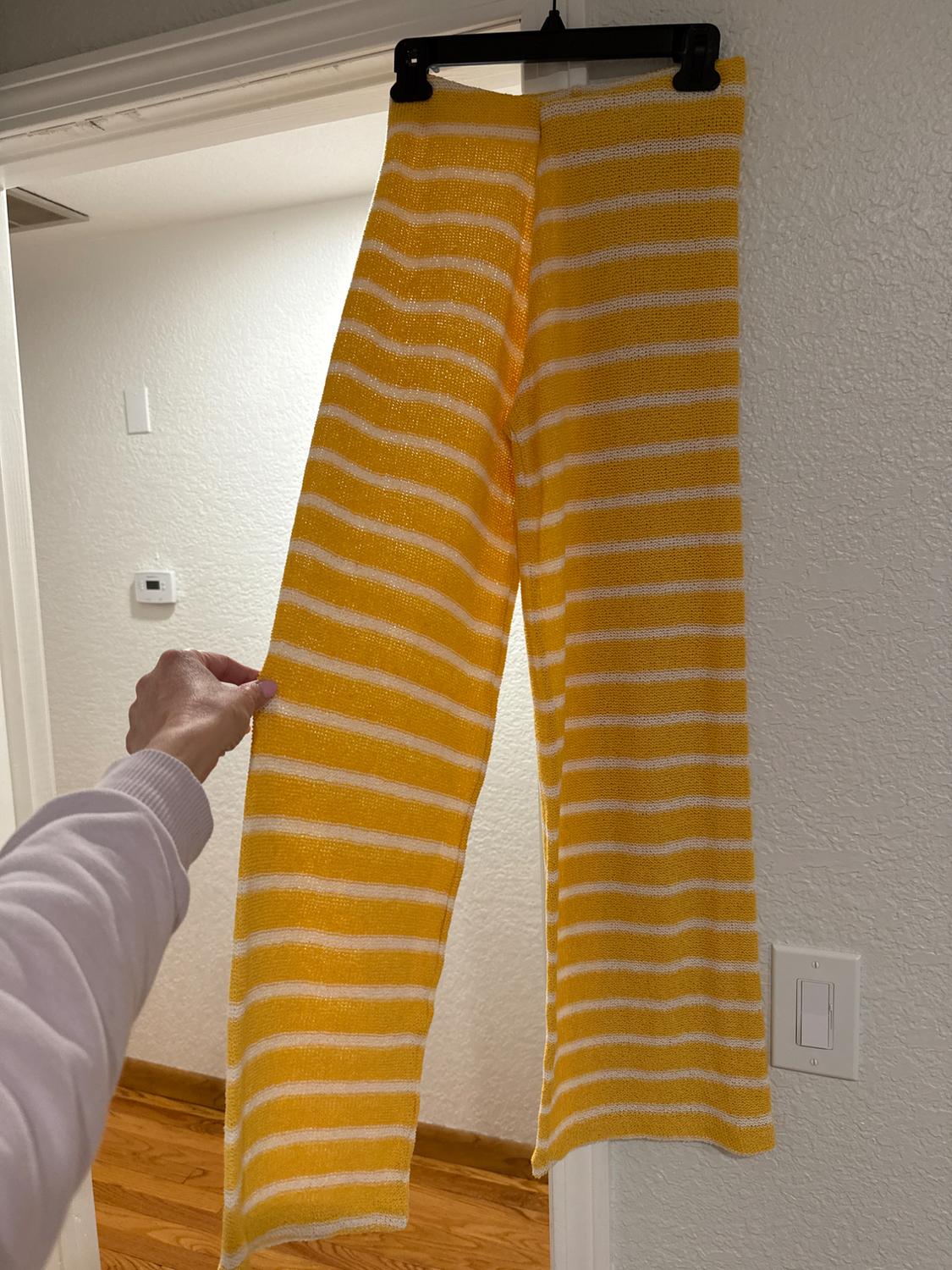 Yellow knit pants with white stripes are hanging on a hanger. A hand is holding the hanger in a well-lit room.