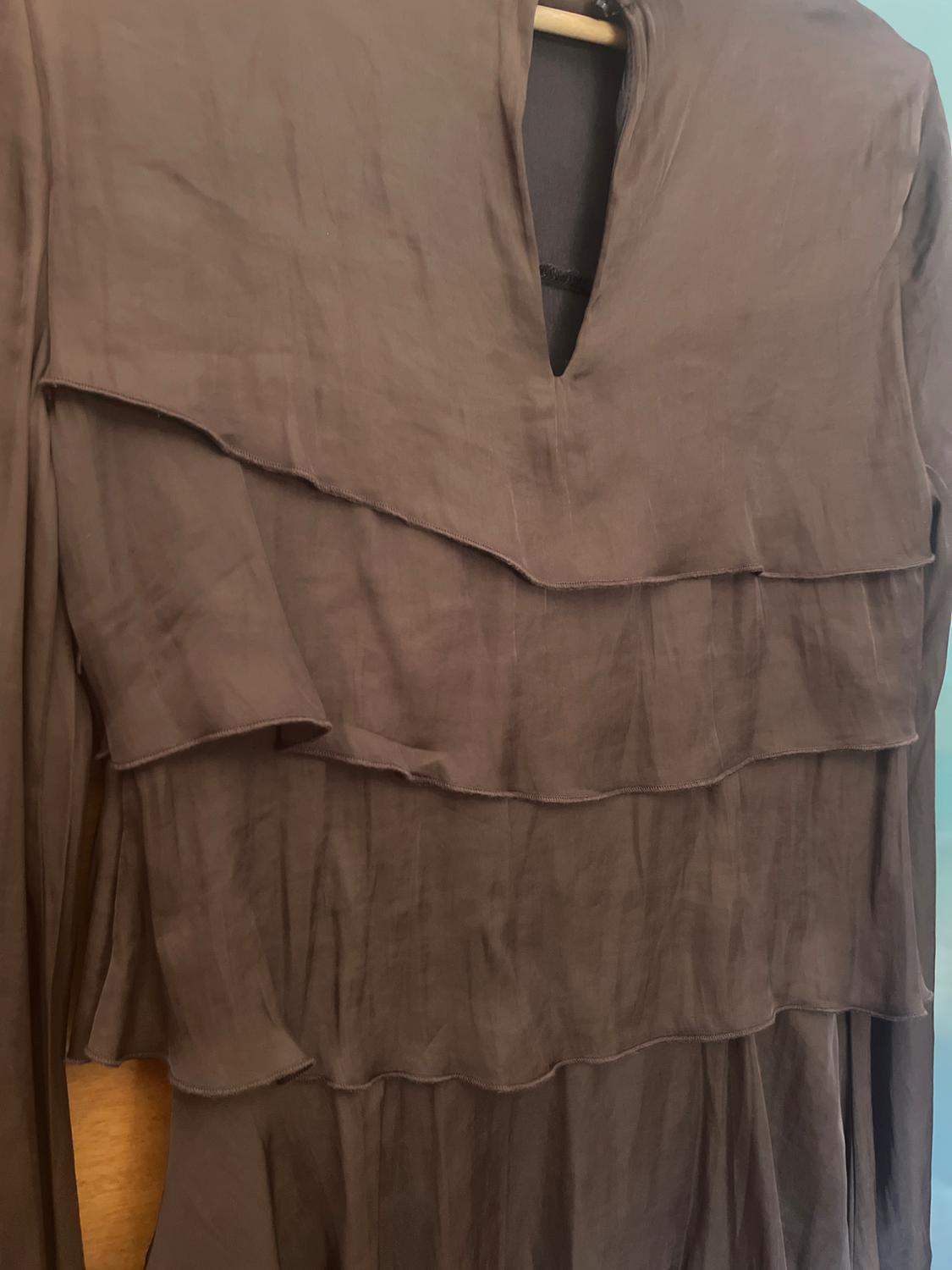 Brown long-sleeved dress with a V-neckline and layered ruffle design, hanging on a wooden hanger in front of a blue wall.