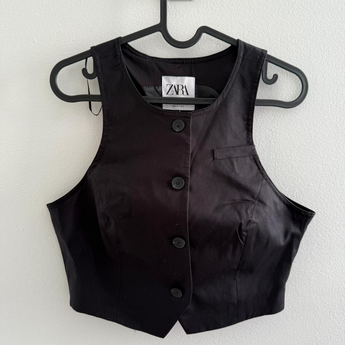 Sleeveless black vest with front buttons and "ZARA" tag, hanging on a black hanger against a white wall.