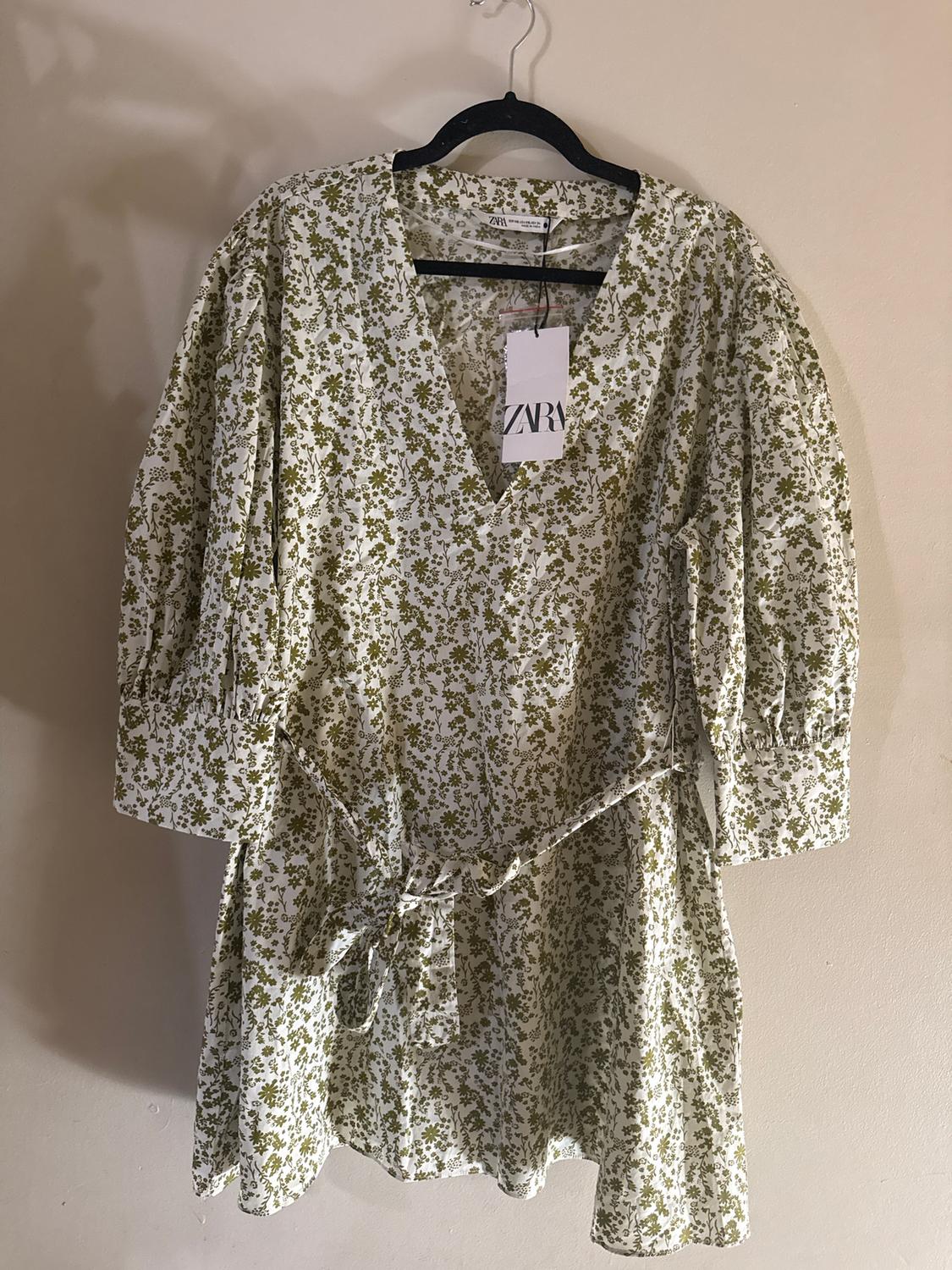 White dress with green floral print, long sleeves, and a belt, hanging on a black hanger. Visible tag with text: "ZARA".